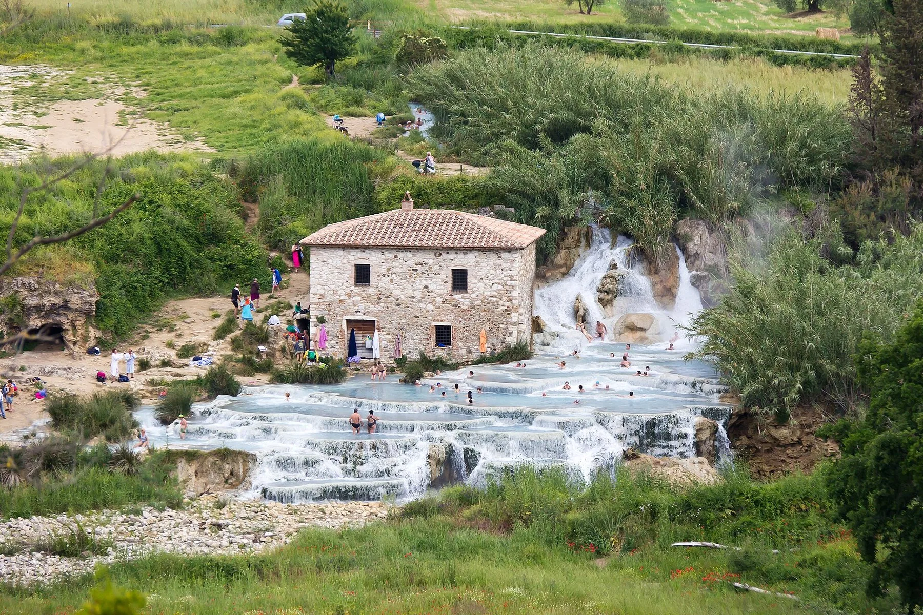 Kalkhvite terrasser med lyseblått vann faller nedover i trappetrinn fra et gammelt steinhus, og badende sitter i de flate bassengene mellom fossene.