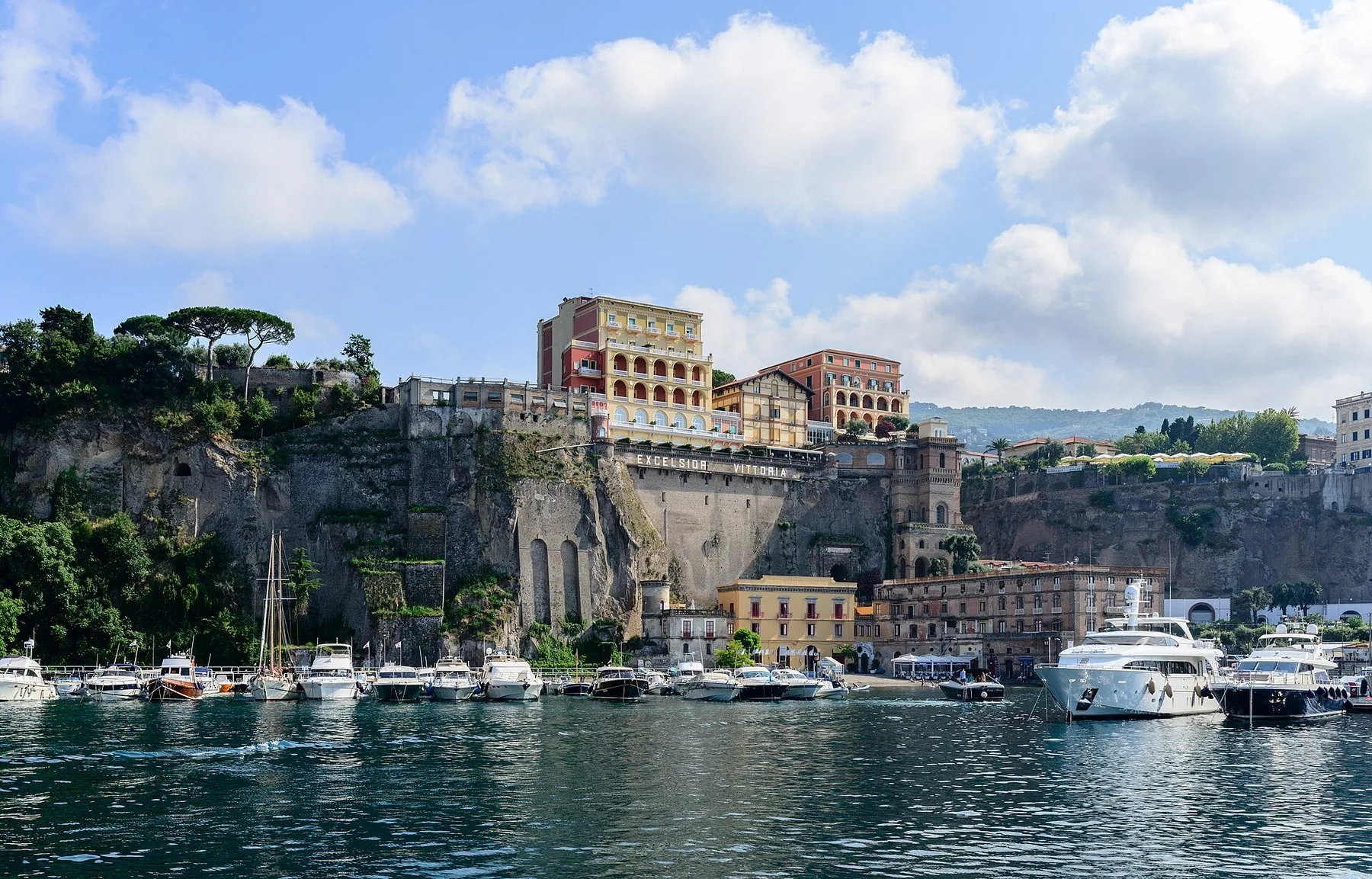 Sorrento ligger på toppen av en loddrett tuffklippe over havna, med hoteller og pinjetrær på kanten og en rekke lystbåter og småbåter langs kaia nedenfor.