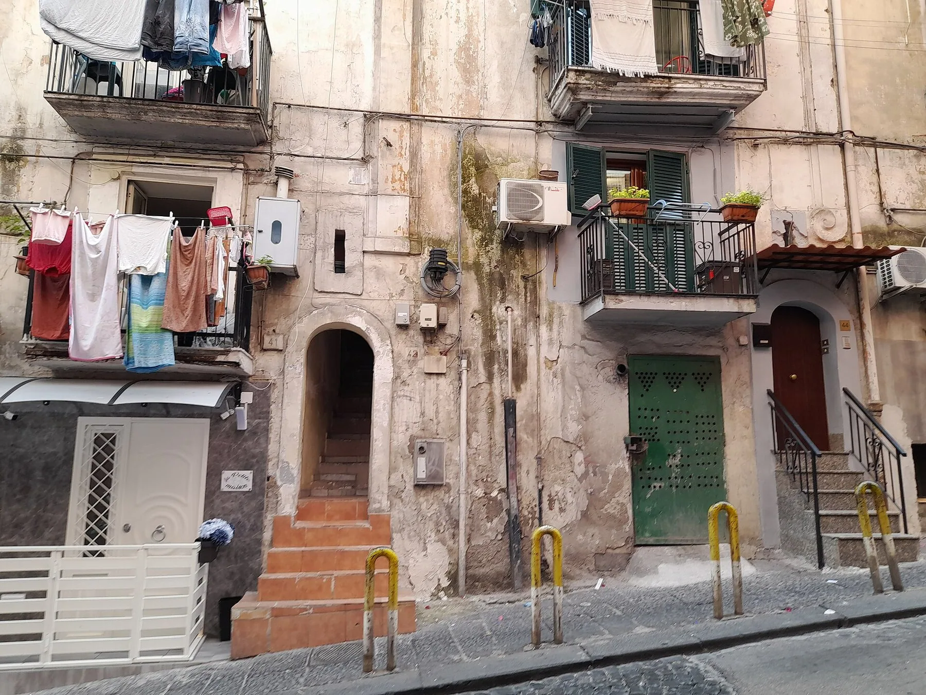 En slitt boligfasade i Quartieri Spagnoli i Napoli med smale balkonger, klesvask hengt til tørk, klimaanleggsvifter på veggen og et hvitt teknisk skap ved inngangstrappen nede på gateplan.