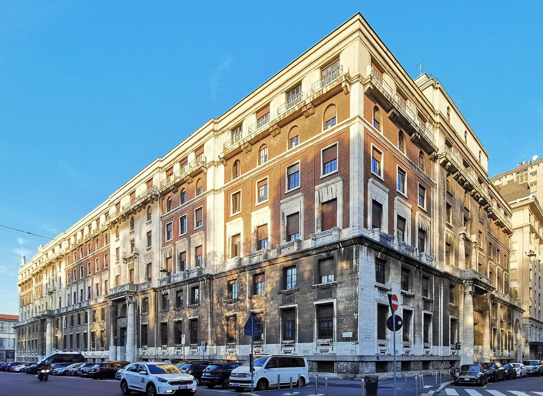 Et stort kontorpalass i lys puss på et gatehjørne i Milano, med avrundet hjørnefasade i fem etasjer, tette vindusrekker og biler parkert langs begge gatene.