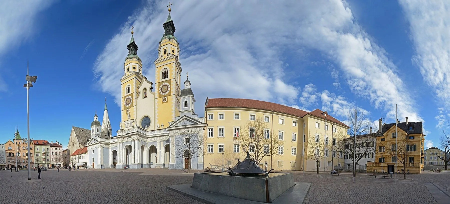 Et bredt torg foran en katedral med to kobbertårn, gule bygninger på siden og fjellsider under blå himmel.