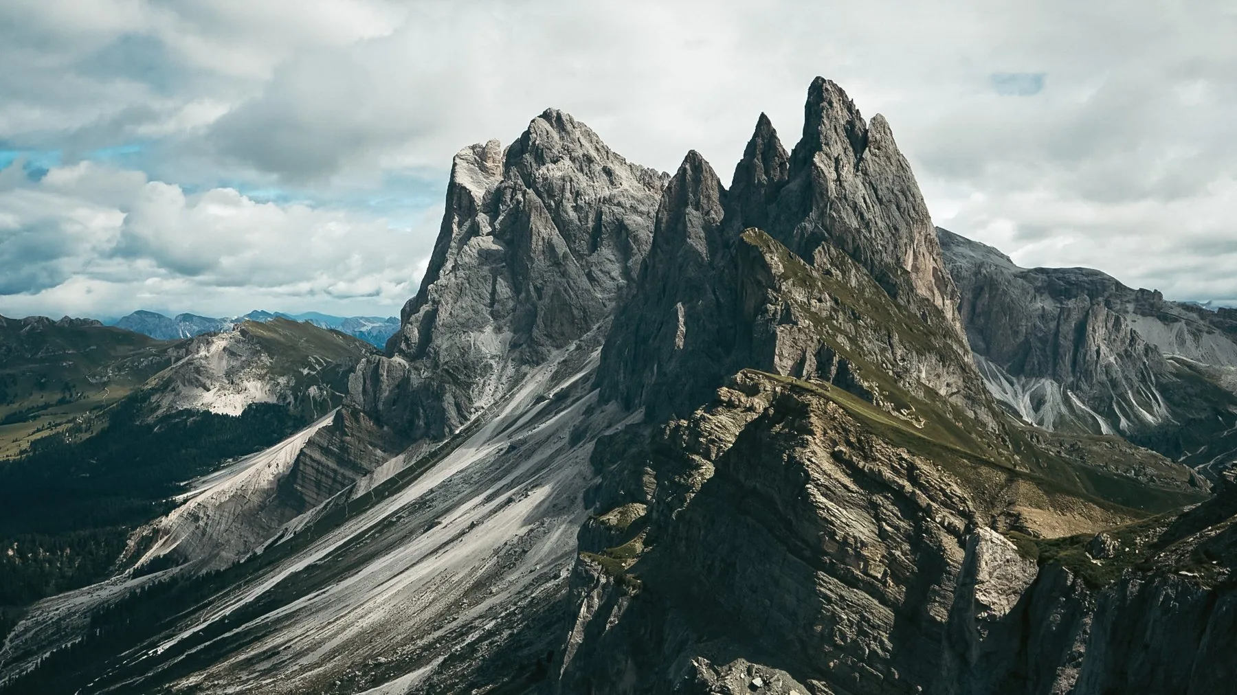Den takkede fjellryggen i Odle-gruppen reiser seg bak grønne alpeenger, med spredte bartrær nederst i lia.