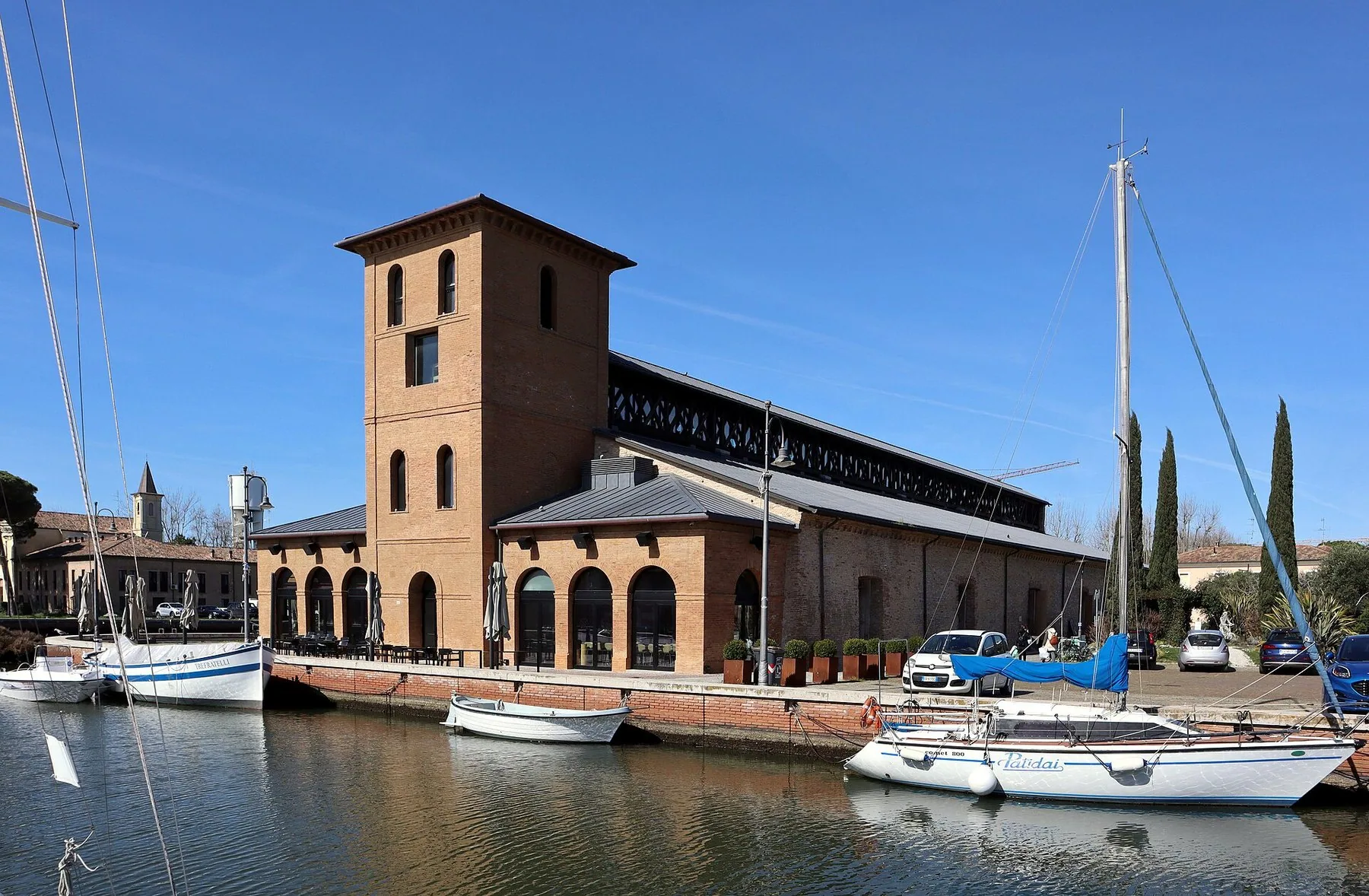 Et gammelt saltlager i rød tegl med tårn ved kanalhavnen i Cervia, med seilbåter fortøyd langs kaia.