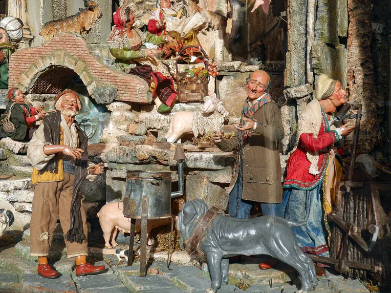 Håndmalte krybbefigurer i gammeldagse klær står tett rundt et bord og en tønne i et miniatyrlandskap av stein og murpuss, med en grå muldyrfigur foran og en hvelvet portåpning bak.