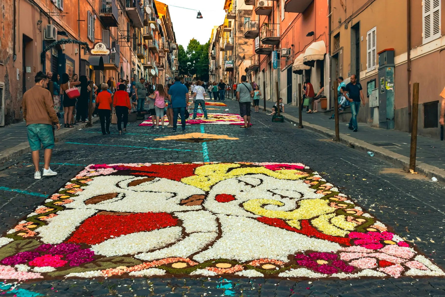 Et teppe lagt av røde, hvite og gule blomsterblader dekker brosteinen i en smal gate, med flere blomsterteppe lenger opp og folk som ser på fra fortauene.