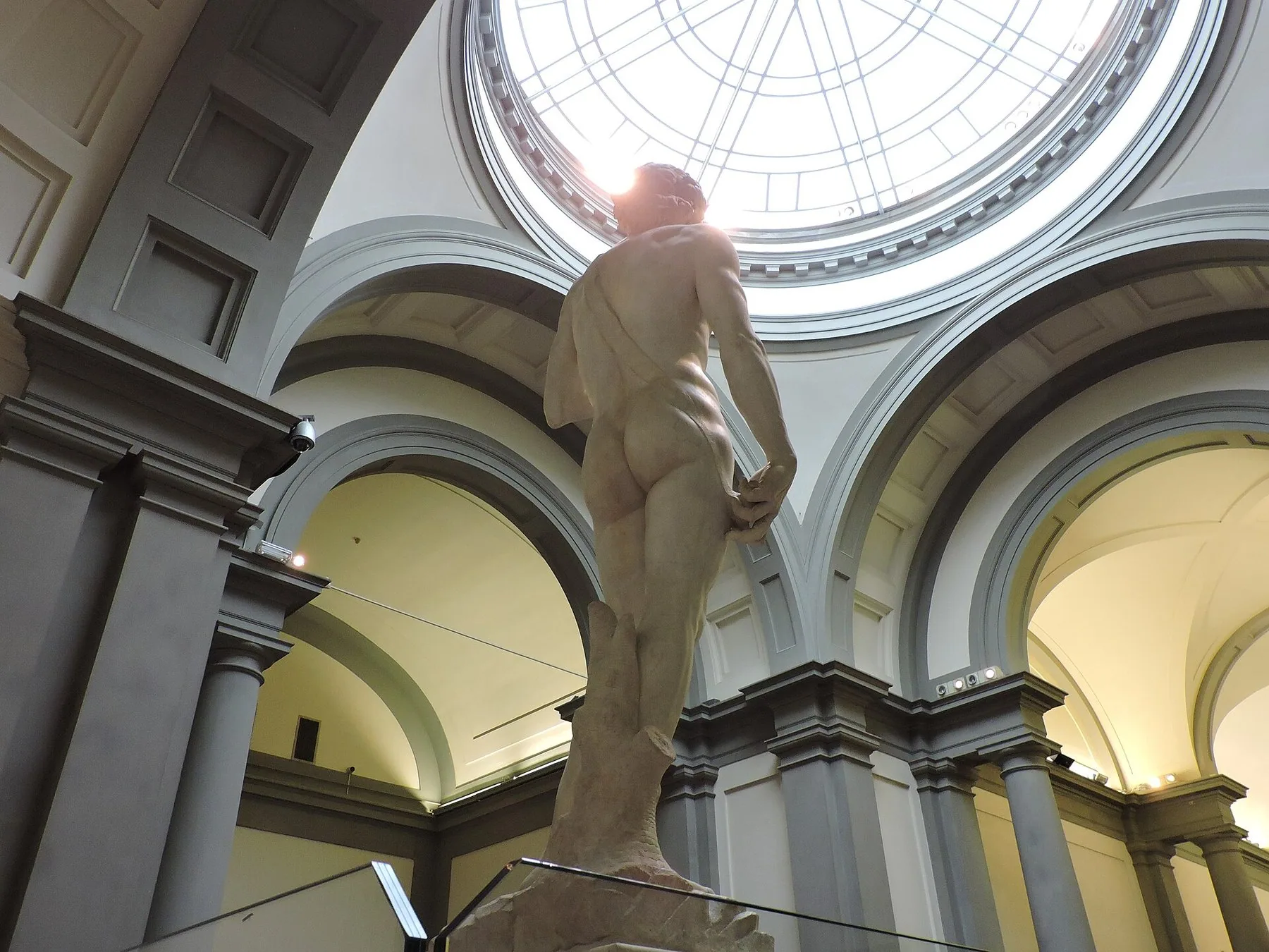 Marmorstatuen av David sett nedenfra i en høy museumssal, med rundbuede hvelv langs sidene og en glasskuppel med overlys rett over figuren.