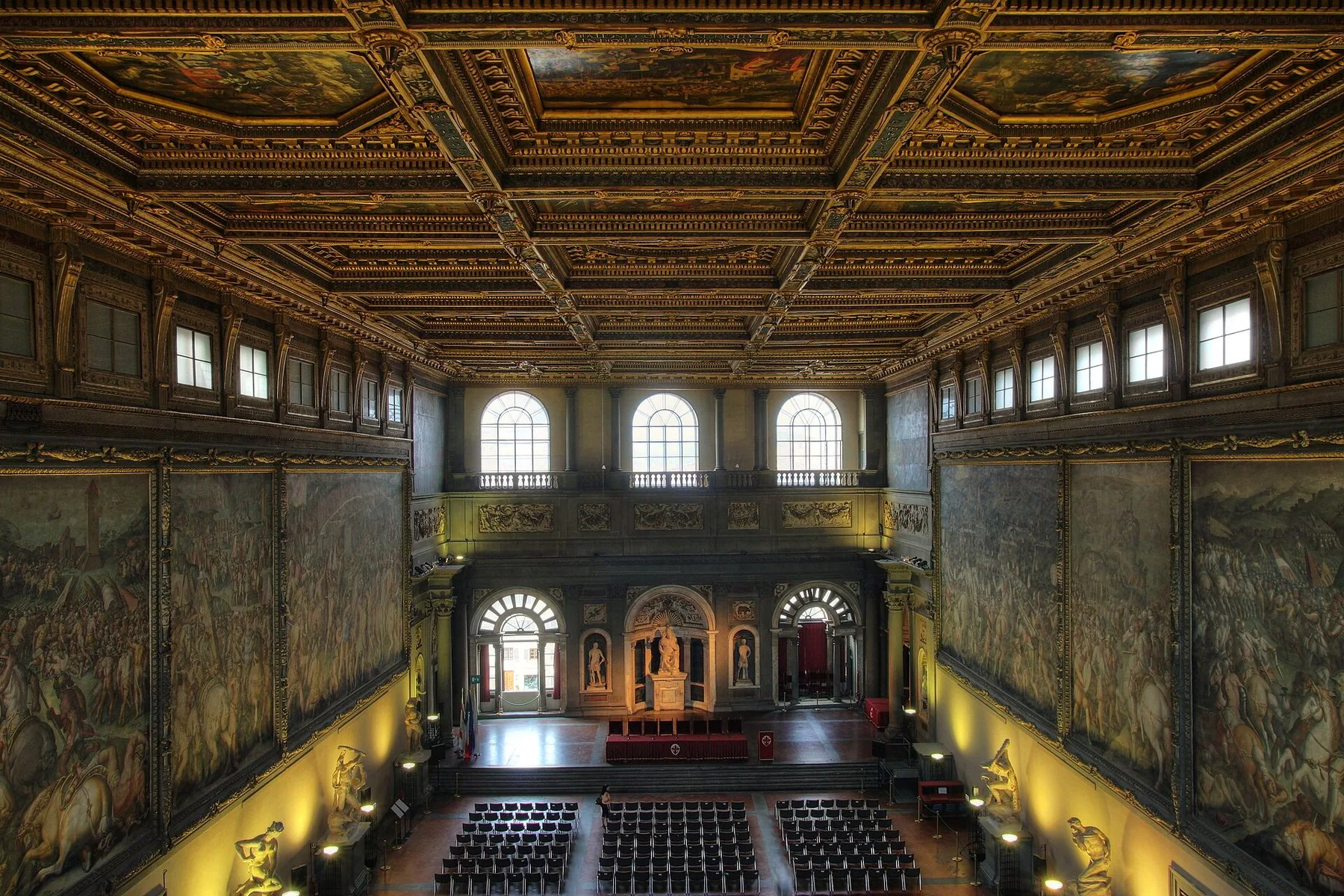 Salone dei Cinquecento i Palazzo Vecchio: en svært høy sal med forgylt kassettak, store slagfresker i full veggbredde på begge sider og rader med stoler nede på gulvet.