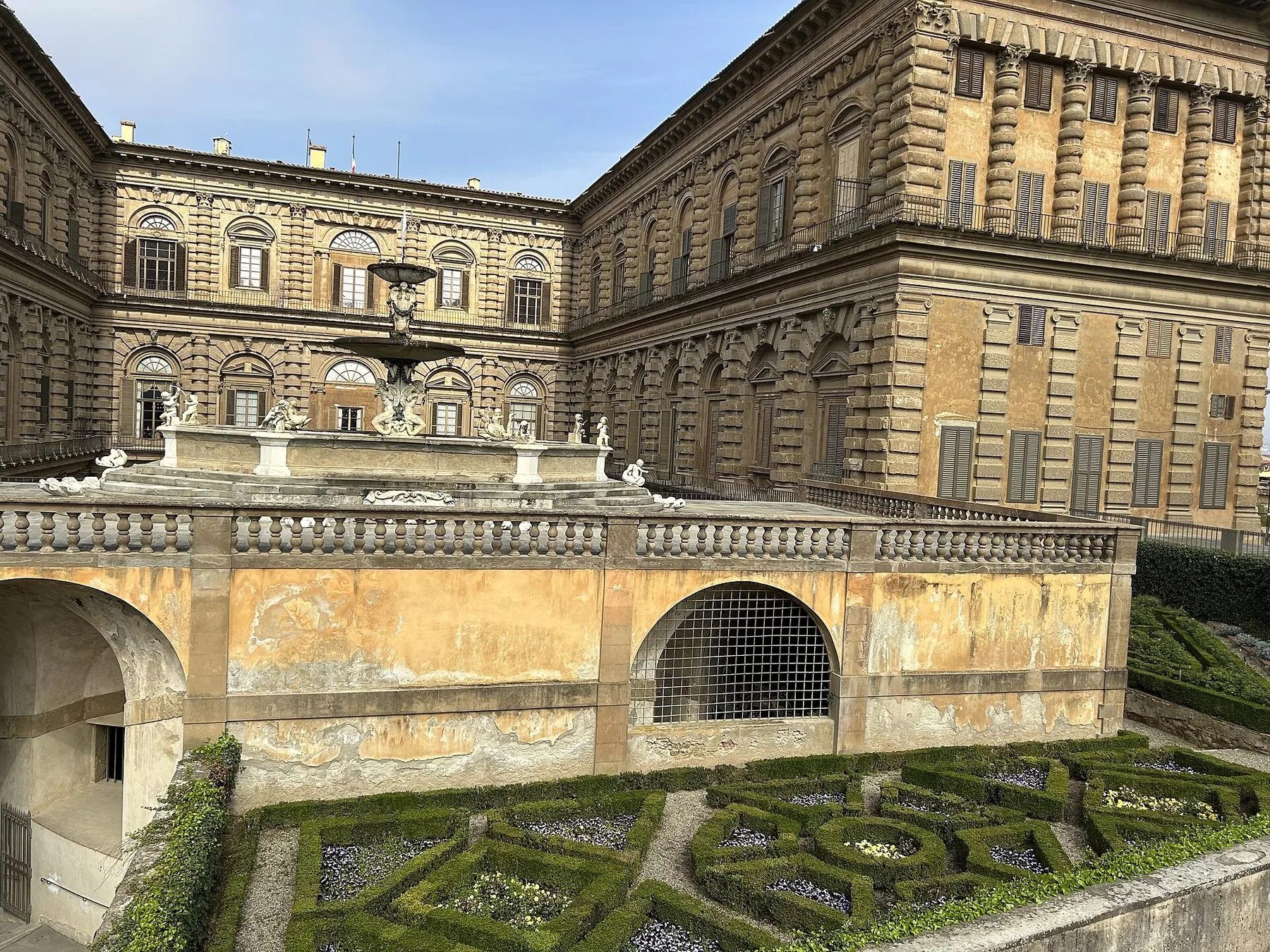 Baksiden av Palazzo Pitti sett fra hagen: den grove natursteinsfasaden i to fløyer, en fontene på terrassen midt i bildet og lave, klipte buksbomsbed nederst i forgrunnen.