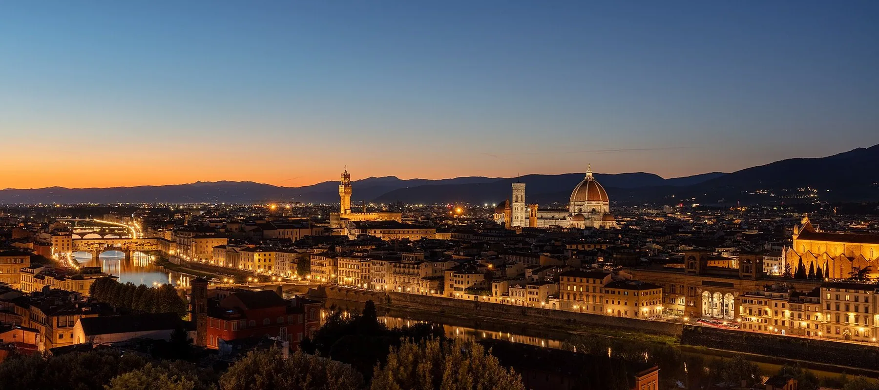 Firenze i skumringen sett fra åssiden i sør: Arno med broene på rekke nedover, det opplyste rådhustårnet midt i bildet og domkuppelen til høyre, mot mørke åser og en oransje horisont.