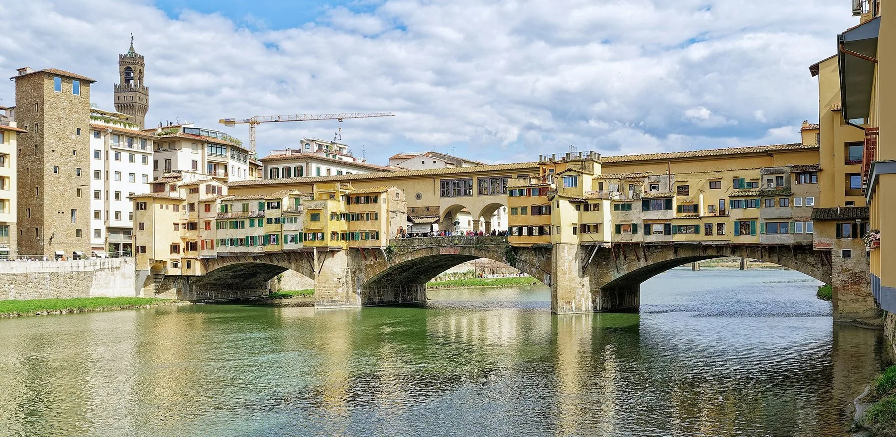 Ponte Vecchio i dagslys sett fra elvebredden: tre steinbuer over Arno og to rekker med små butikklokaler som henger utover vannet på utkragede bjelker, med et høyt bytårn bak til venstre.