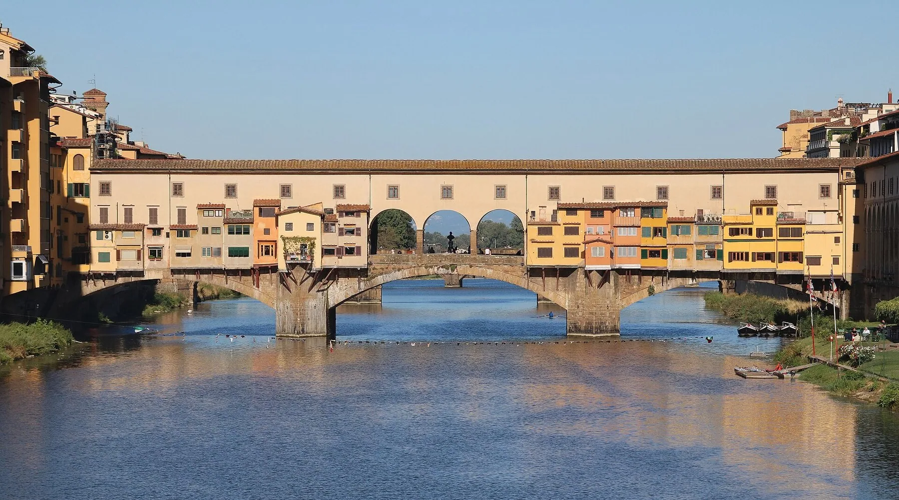 Ponte Vecchio med de gule butikkene som henger ut over elven, speilet i det stille vannet, og husrekker i oker og rosa på begge bredder.