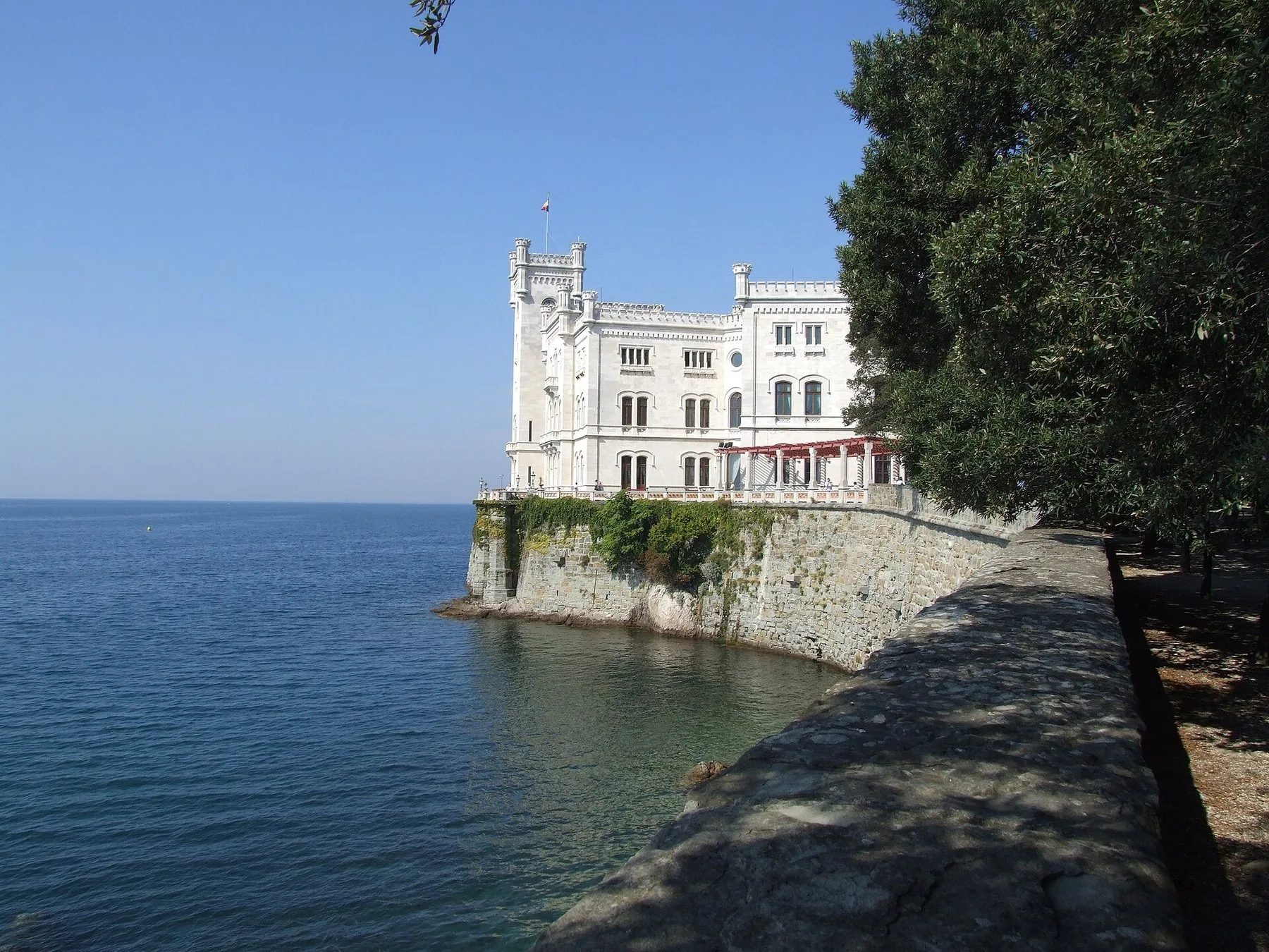 Det hvite slottet Miramare ved Trieste står ytterst på en steinpynt i havet, sett fra en mur i skyggen av store trær.