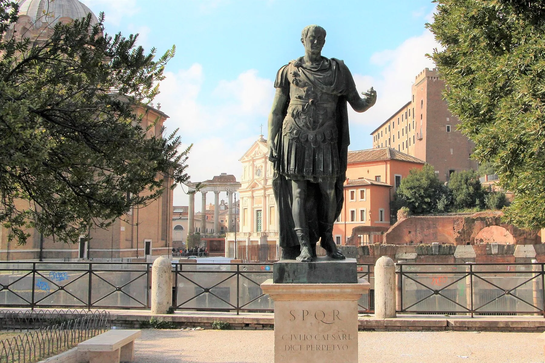 Bronsestatue av Julius Cæsar i rustning på en sokkel med innskriften SPQR, med pinjetrær og fargerike bygninger i bakgrunnen.