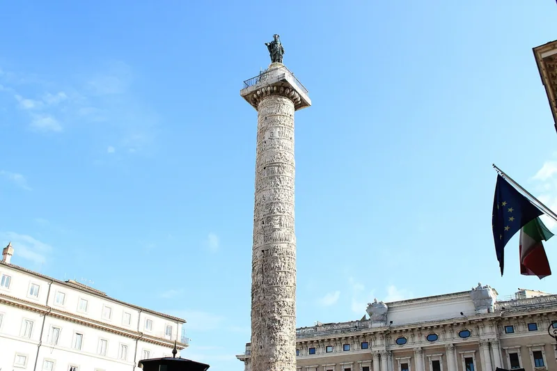 Marcus Aurelius-søylen med spiralrelieffer og en bronsestatue på toppen, mot blå himmel med flagg og palassfasader nedenfor.