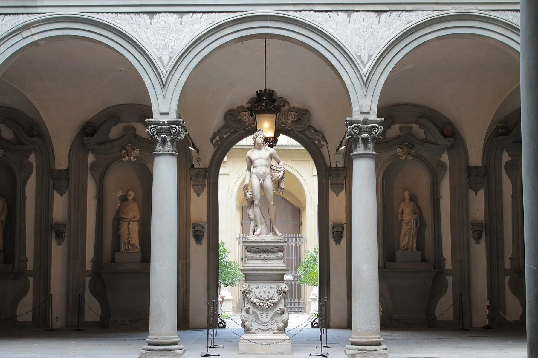 Et kvadratisk borggårdsrom med hvite søyler og rundbuer på alle sider, en marmorstatue i nisjen midt imot og mindre figurer i nisjene til side.