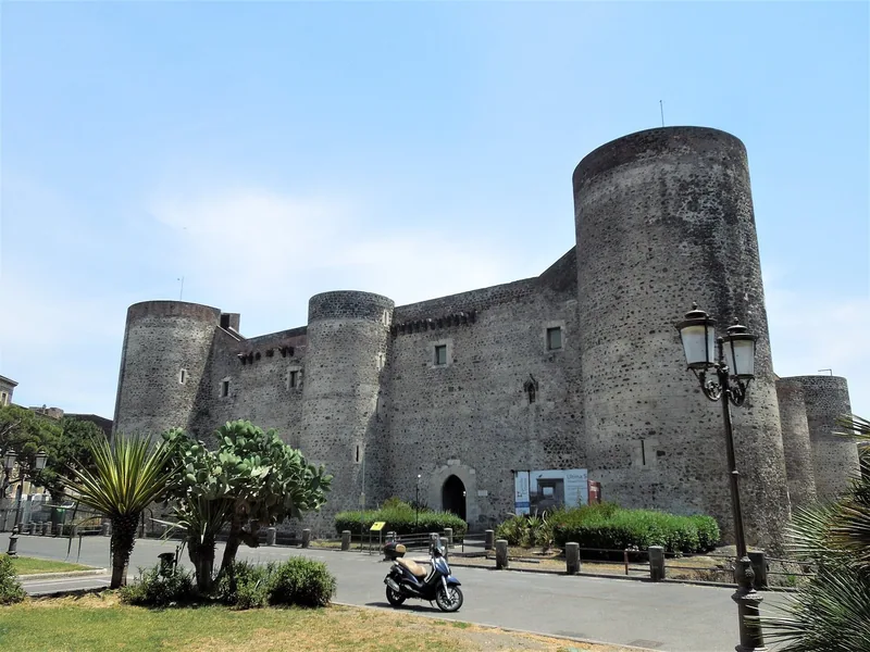 Castello Ursino: en firkantet borg av mørk lavastein med runde hjørnetårn, sett fra en plass med palmer, kaktus og en gatelykt.
