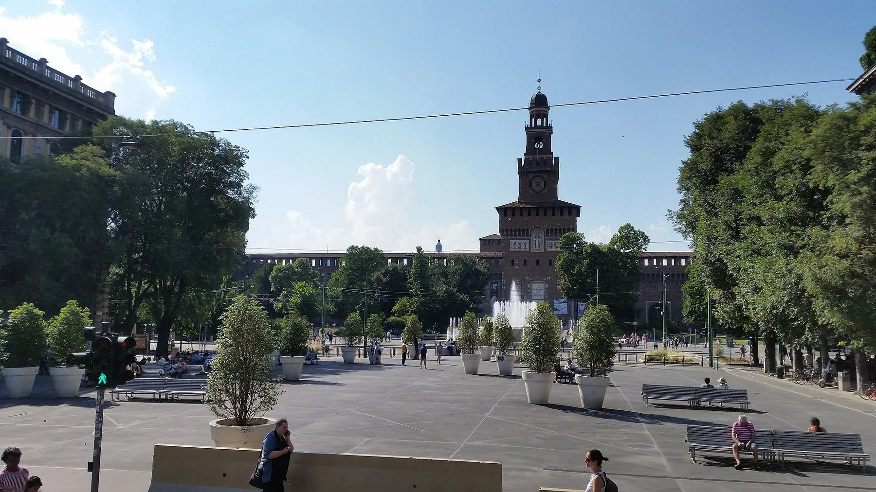 Castello Sforzesco i Milano med det høye inngangstårnet midt på teglfasaden, en fontene og en åpen plass med trær foran.