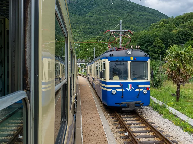 Et blått og hvitt smalsportog kjører inn ved en liten stasjon i en grønn fjelldal, sett fra vinduet på et annet tog.