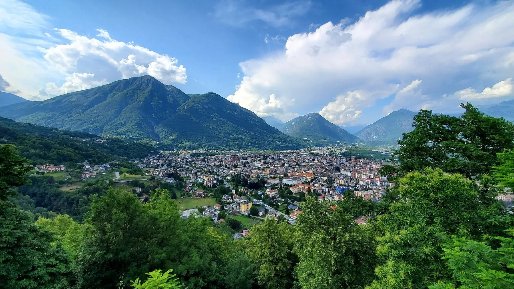 Domodossola ligger i en vid dalbunn omgitt av grønne fjell, sett fra en skogkledd skråning en klar dag.