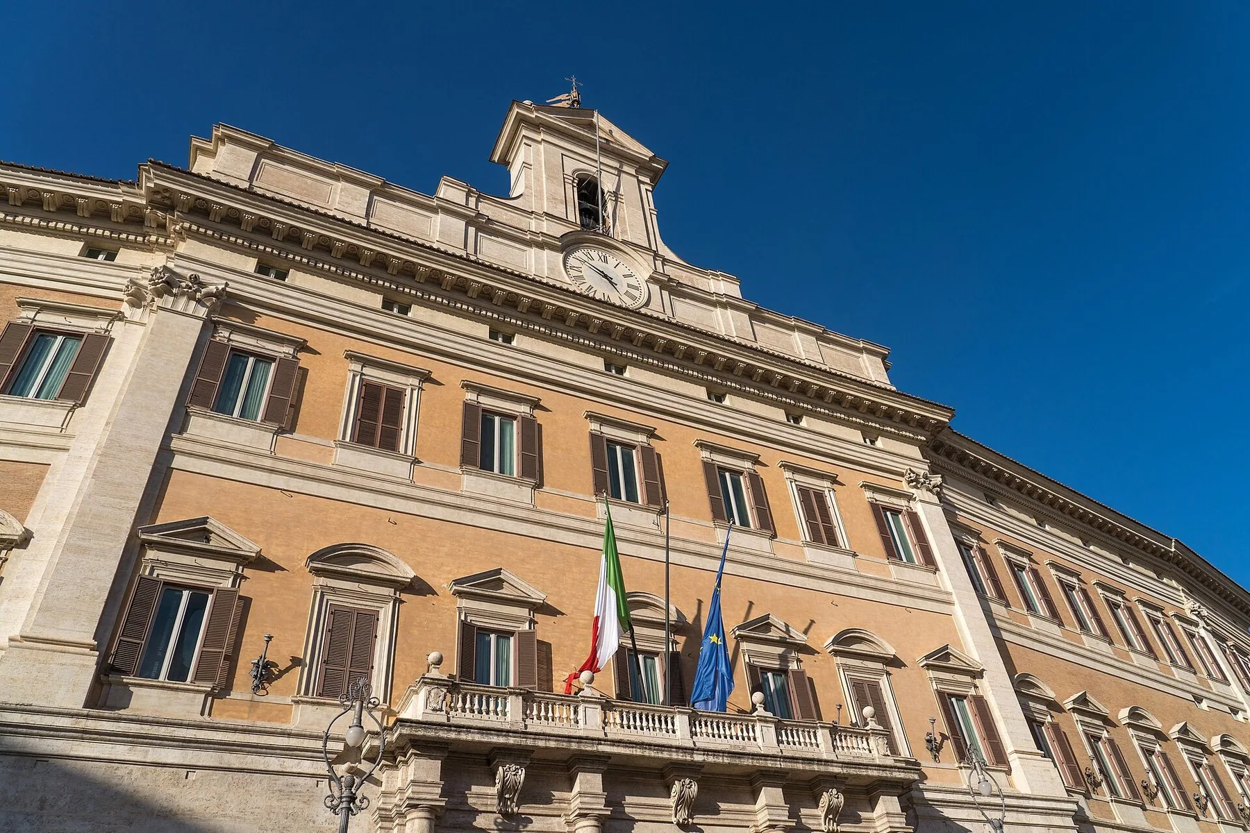 Fasaden på Palazzo Montecitorio i Roma, der Deputertkammeret holder til, sett nedenfra med klokketårn, balkong og italiensk og europeisk flagg mot dypblå himmel.