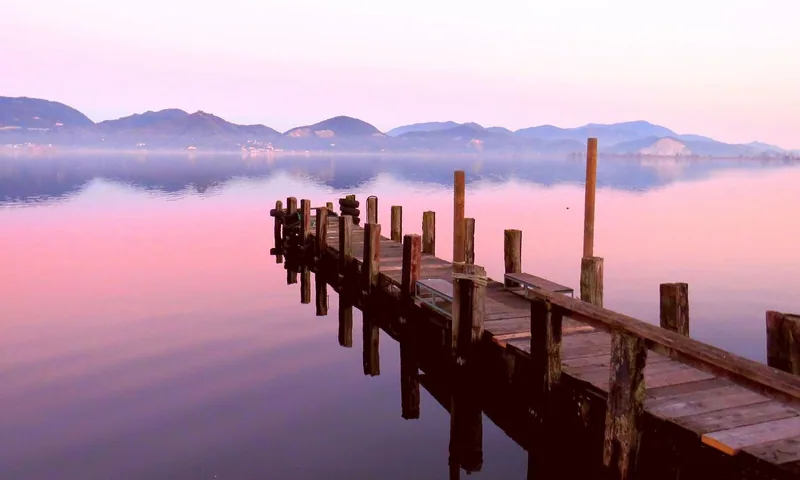 En trebrygge går ut i Massaciuccoli-sjøen ved Torre del Lago i rosa morgenlys, med fjell som speiler seg i det stille vannet.