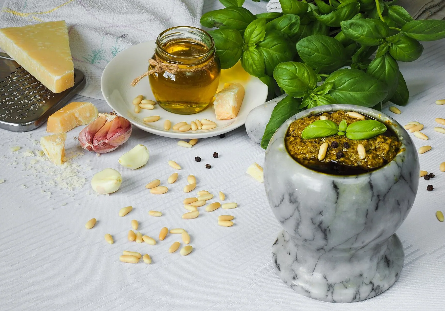 En marmormorter fylt med grønn pesto, omgitt av basilikum, pinjekjerner, hvitløk, revet ost og en glasskrukke olivenolje.