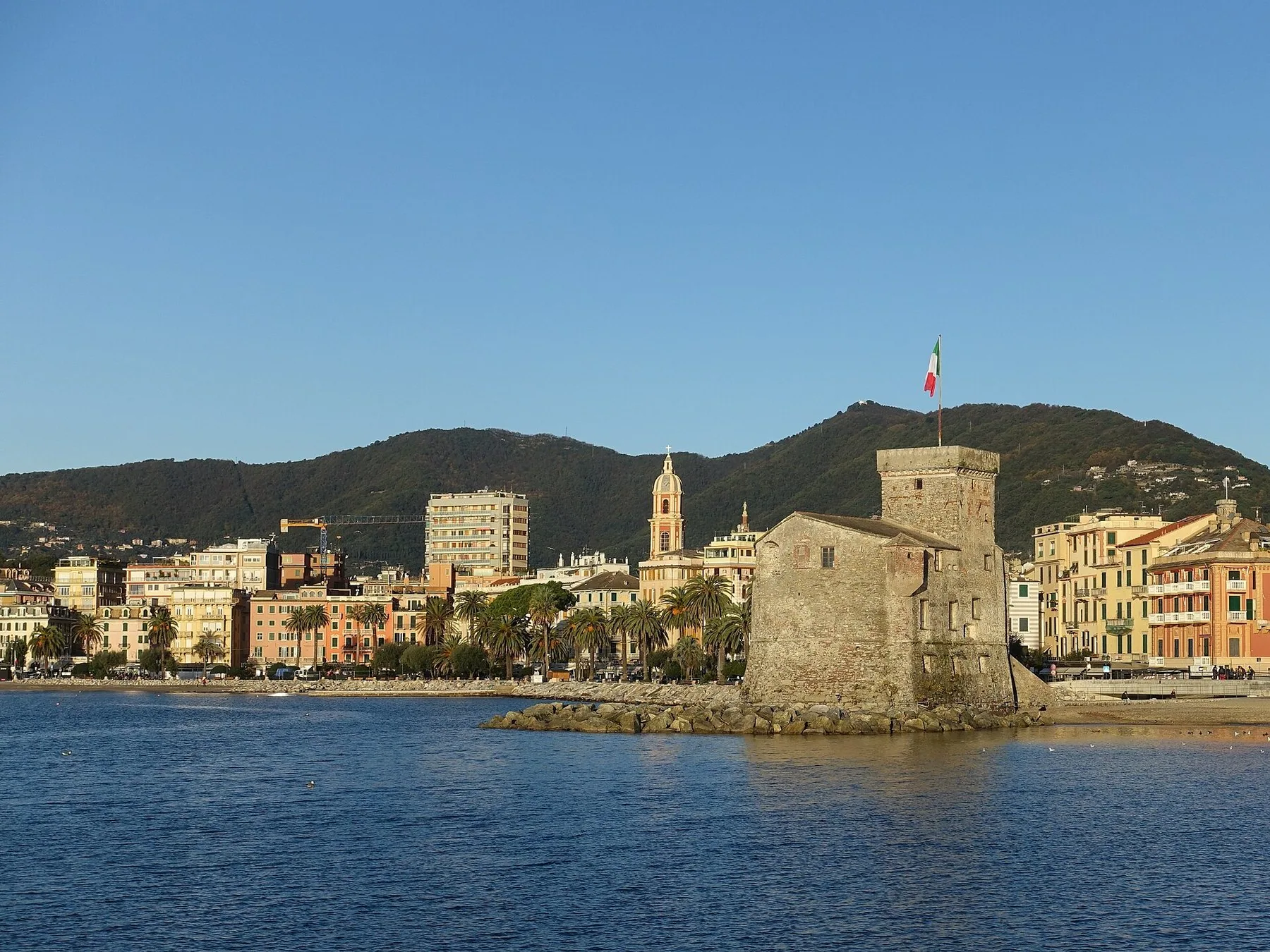 Det lille slottet på moloen i Rapallo med italiensk flagg, foran strandpromenaden og grønne åser.