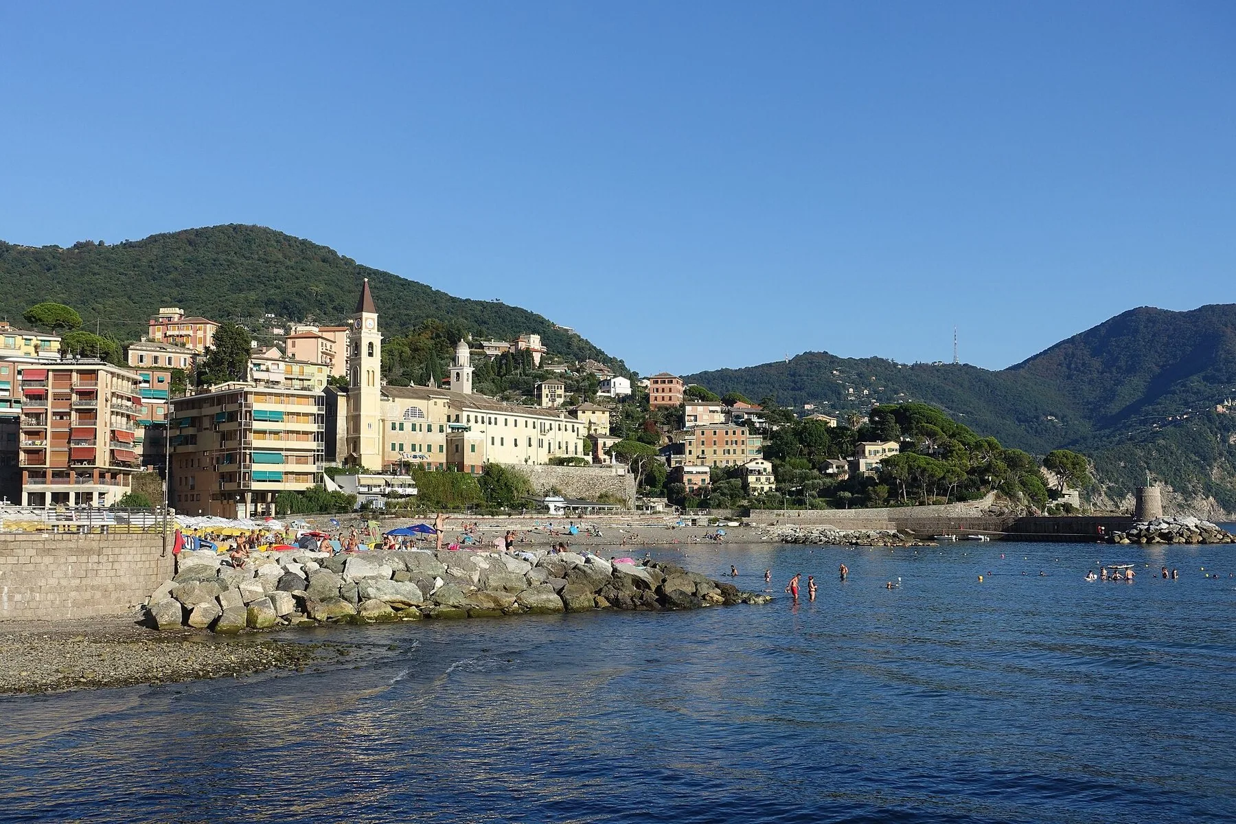 Strandpromenaden i Recco med fargerike hus, et kirketårn og grønne åser bak, og en steinmolo ut i det rolige havet.