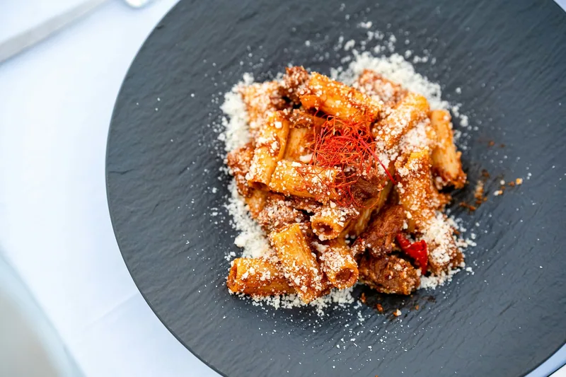Rigatoni i rød tomatsaus med kjøttbiter, dryppet over med revet hvit ost og tynne tråder av tørket chili, servert på en mørk skifertallerken.