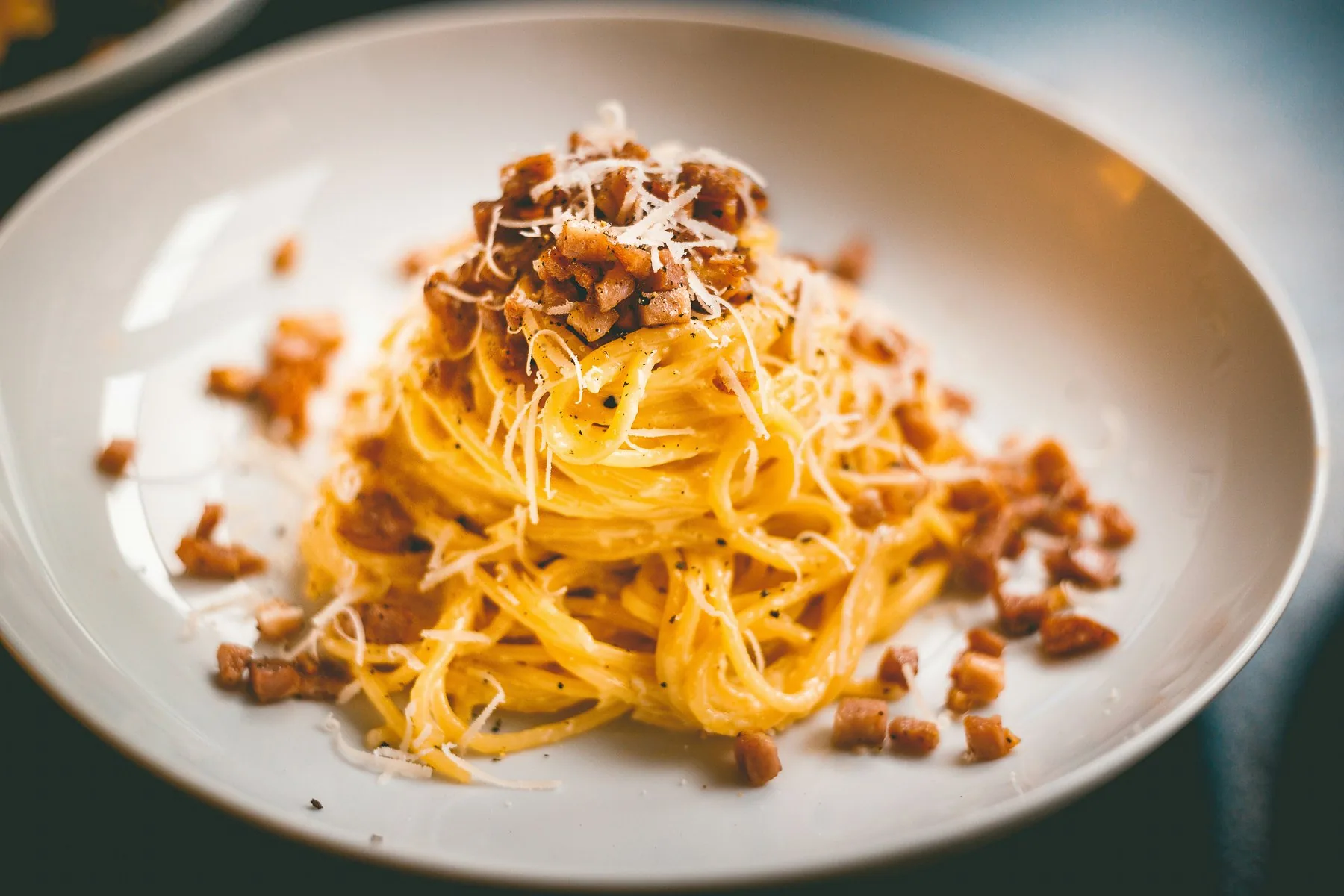 Spaghetti vendt i en glinsende, gul eggesaus, toppet med sprøstekte terninger av svinekjøtt og revet ost i en dyp, hvit tallerken.