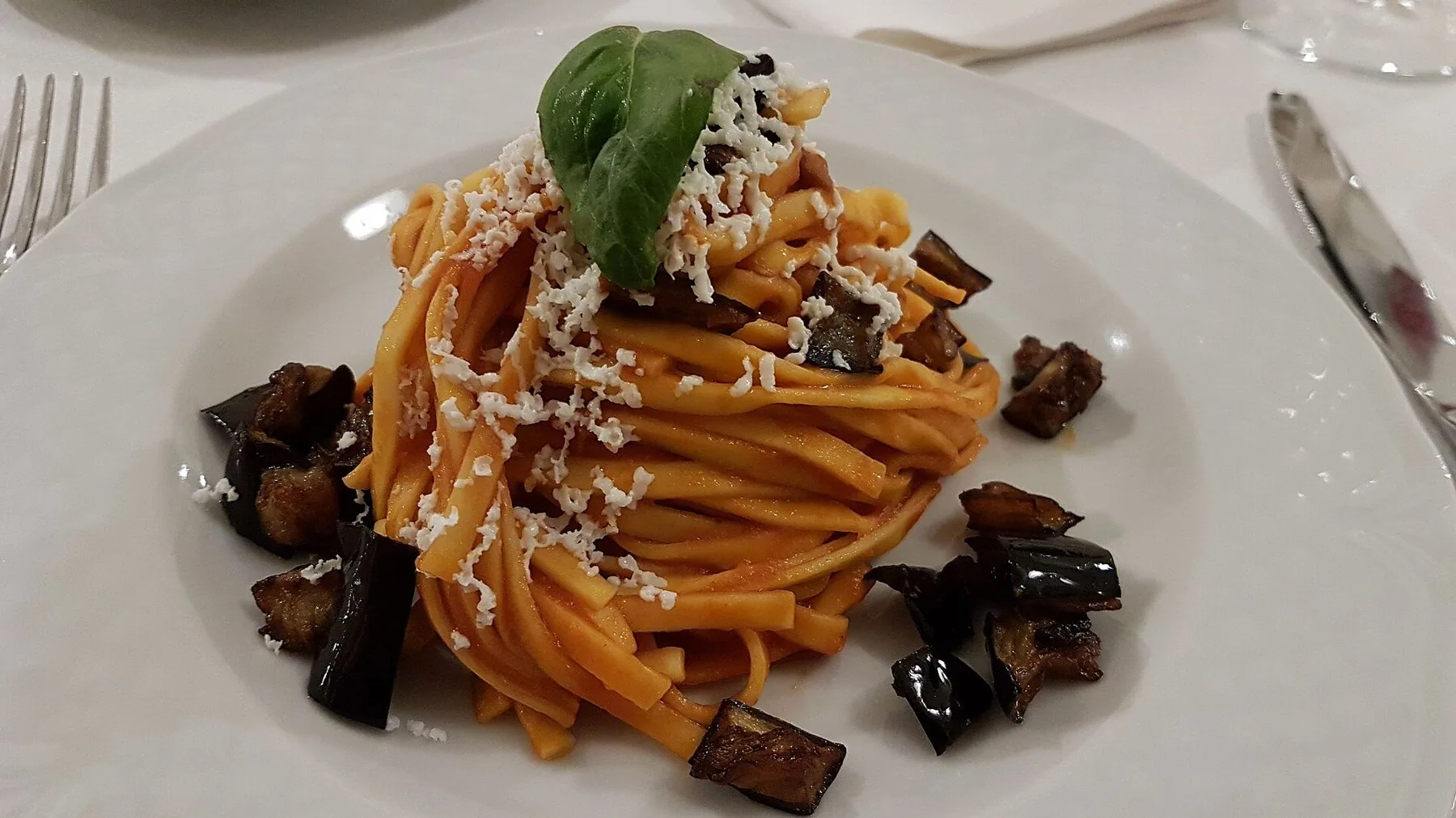 Pasta alla Norma på hvit tallerken: tomatsaus, stekte aubergineterninger, revet ricotta salata og et basilikumblad på toppen.