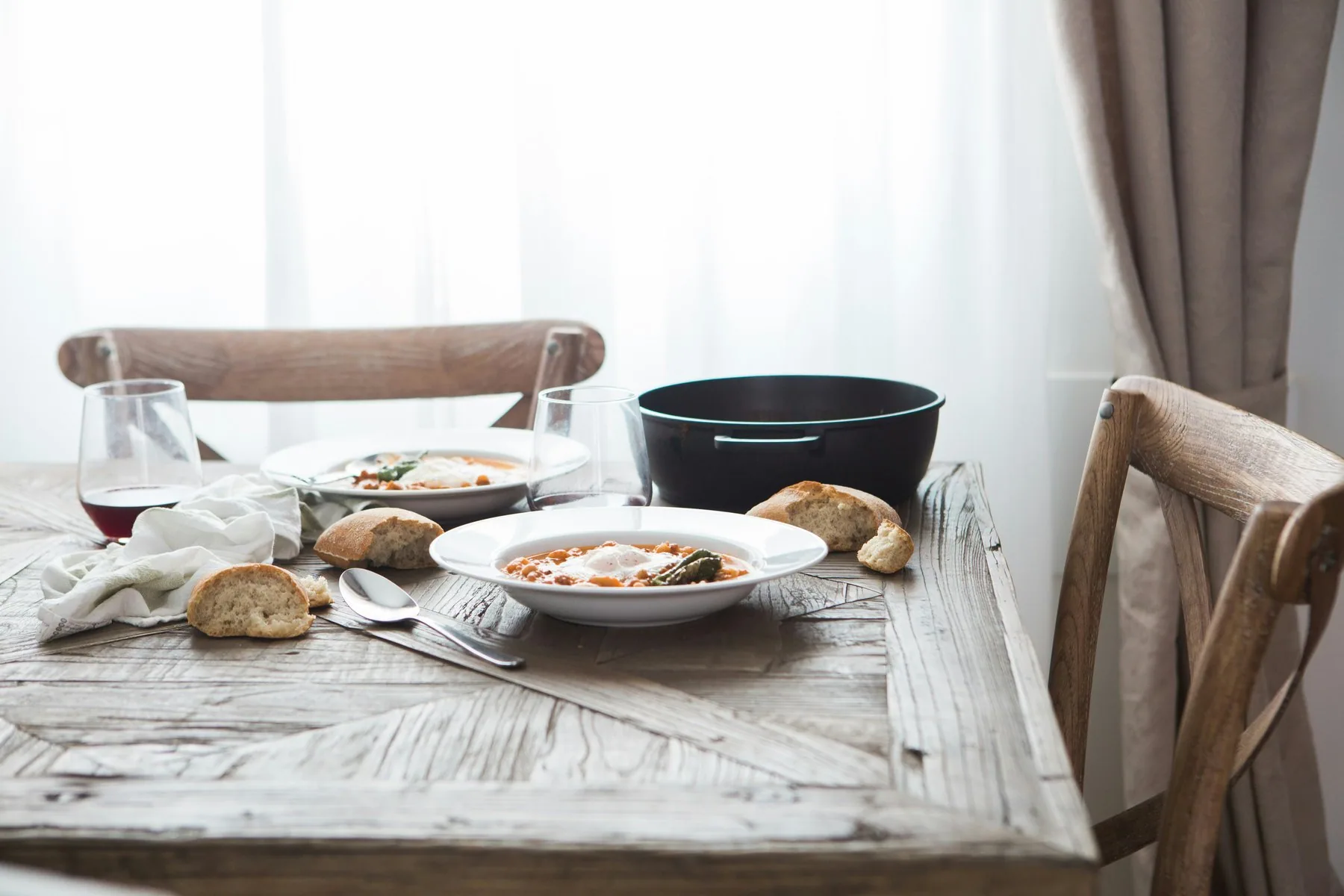 To dype hvite tallerkener med en tykk bønnesuppe står på et grått trebord sammen med brutte brødstykker, en skje, et glass rodvin og en svart gryte i bakgrunnen.