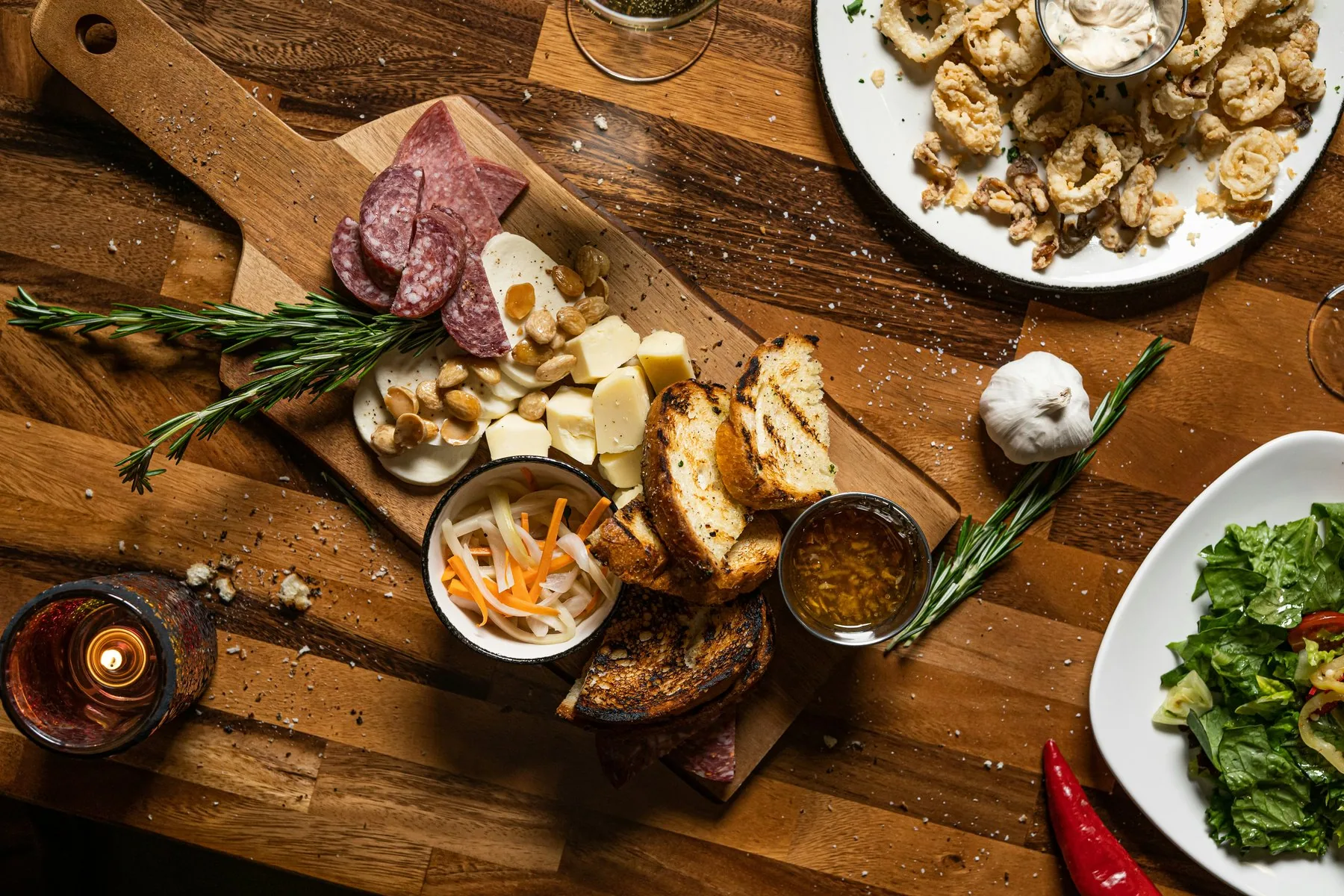 Et trefjøl på et bord med salamiskiver, ostebiter, ristet brød og små skåler med syltede grønnsaker, med en tallerken fritert sjømat og en salatbolle ved siden av.