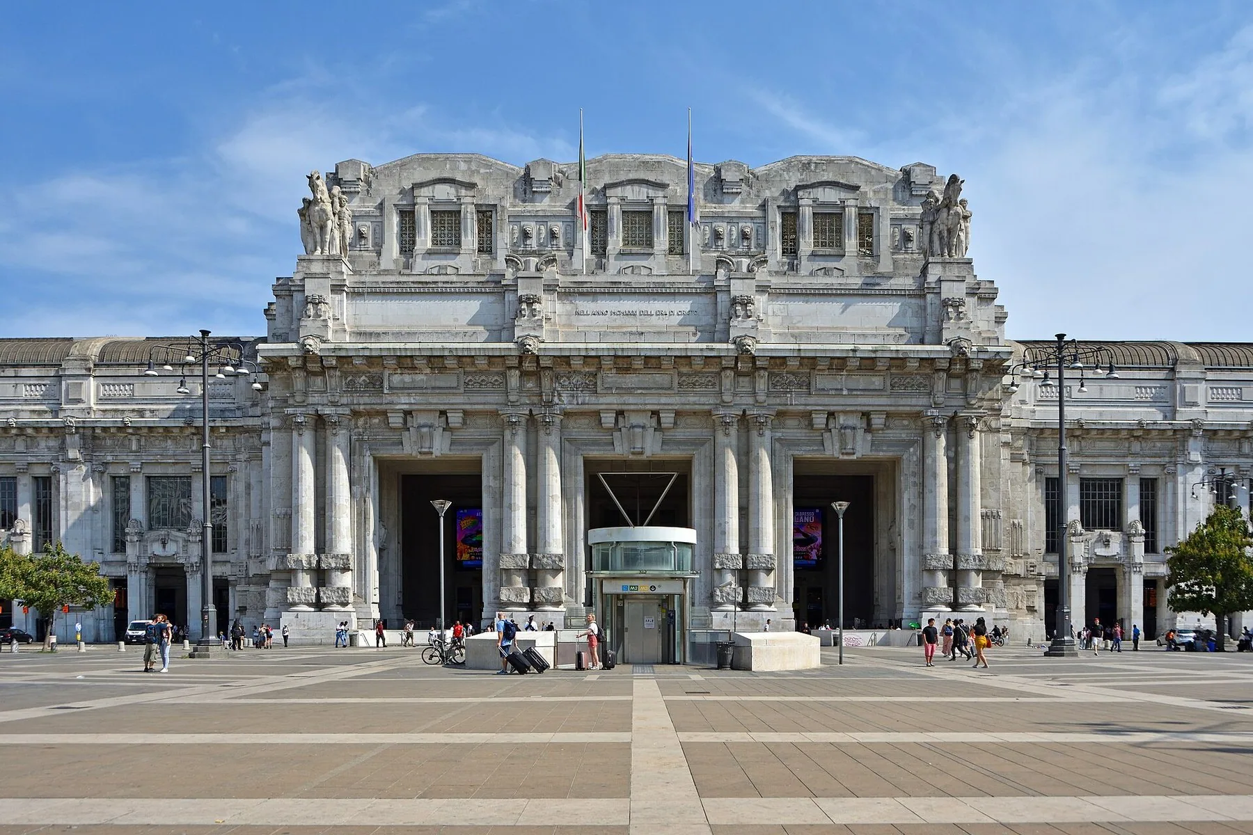 Den monumentale steinfasaden på Milano Centrale, med søyler, skulpturer og høye buede innganger bak en bred, nesten tom plass.