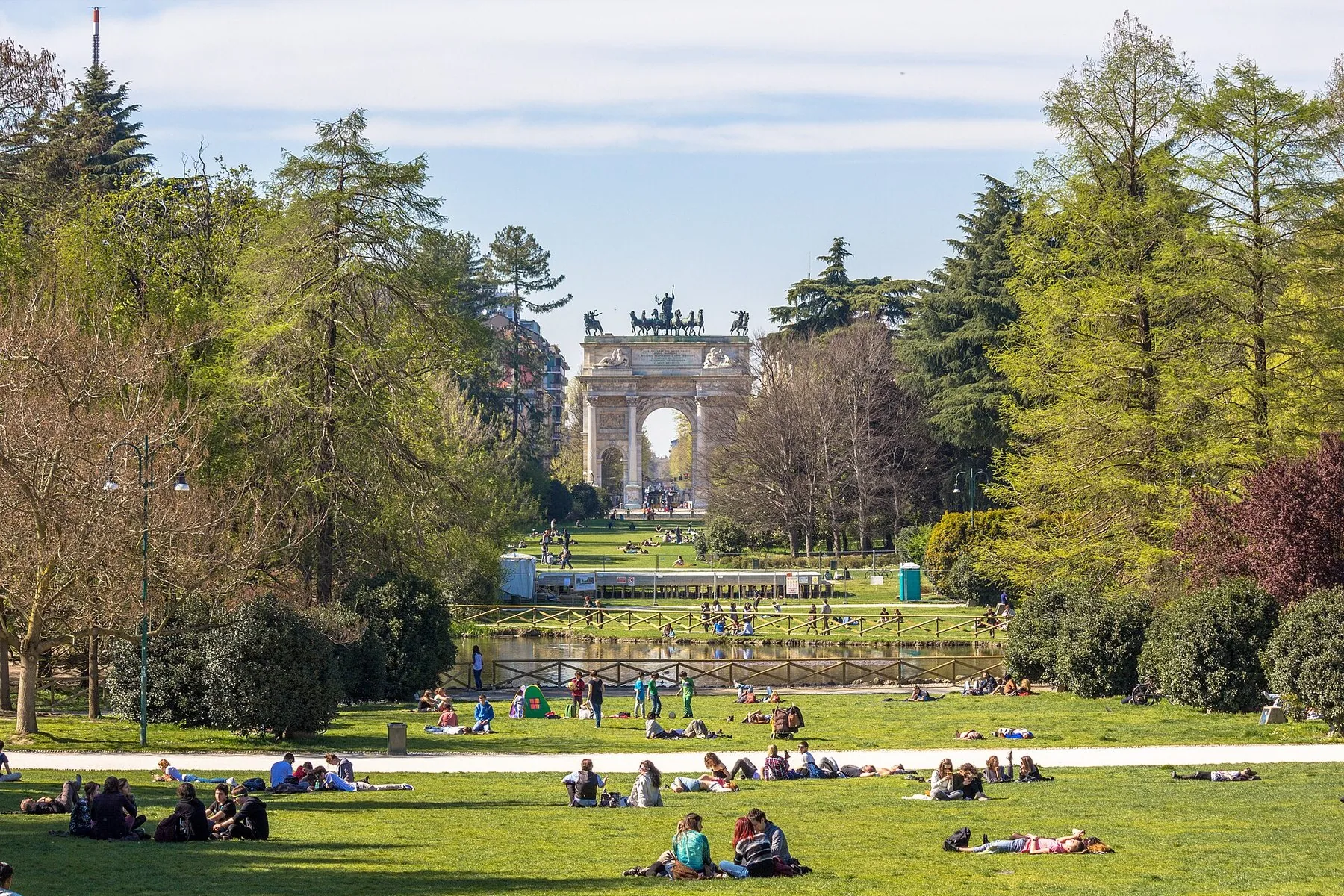 En stor gressflate i Parco Sempione der folk sitter og ligger spredt utover, med høye løvtrær langs begge sider og en triumfbue i stein i enden av siktlinjen.