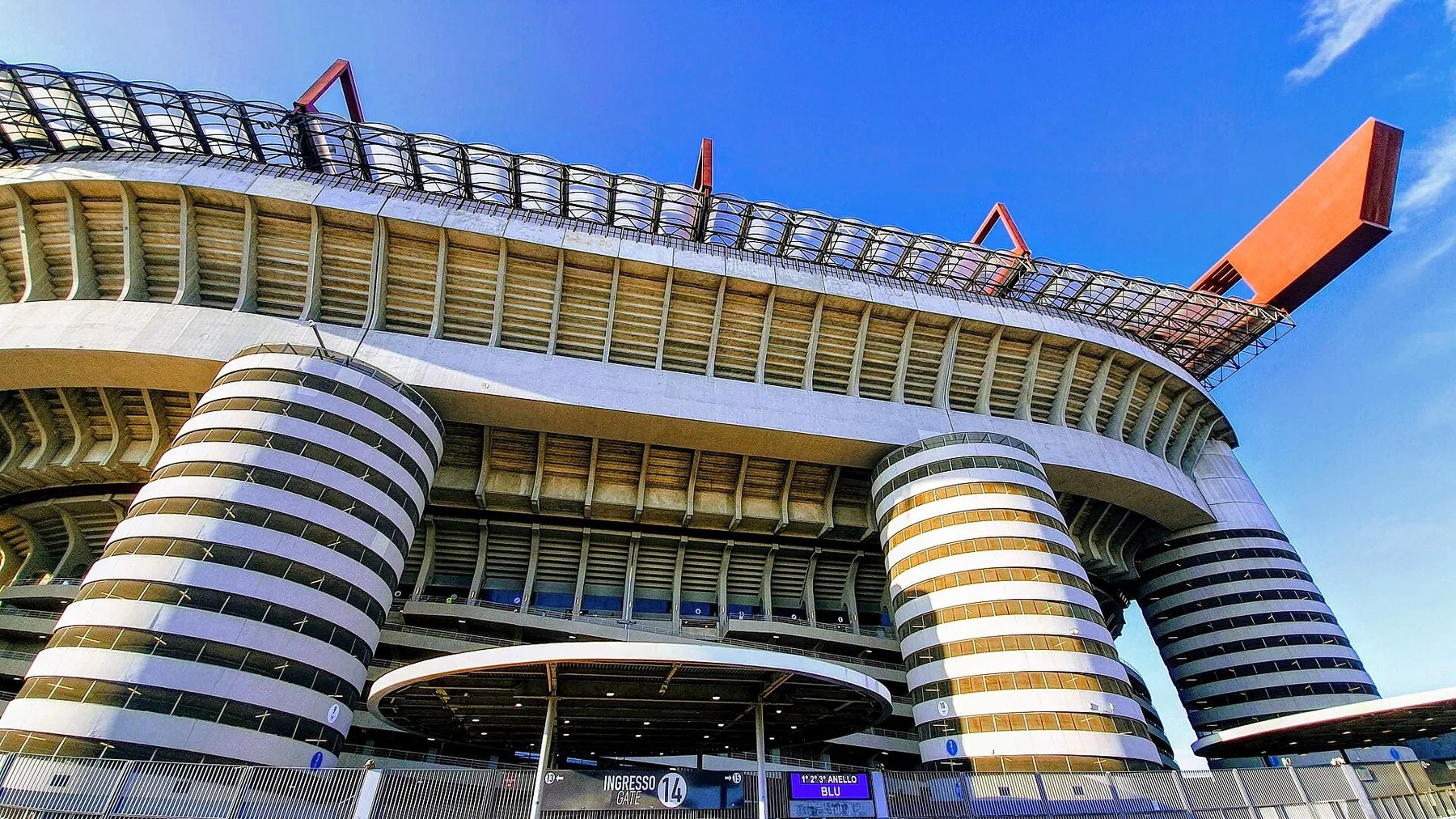 Stadion på San Siro sett nedenfra: de sylinderformede spiralrampene i betong, tribuneetasjene som hviler på dem og de røde takdragerne som stikker ut mot en klar blå himmel.