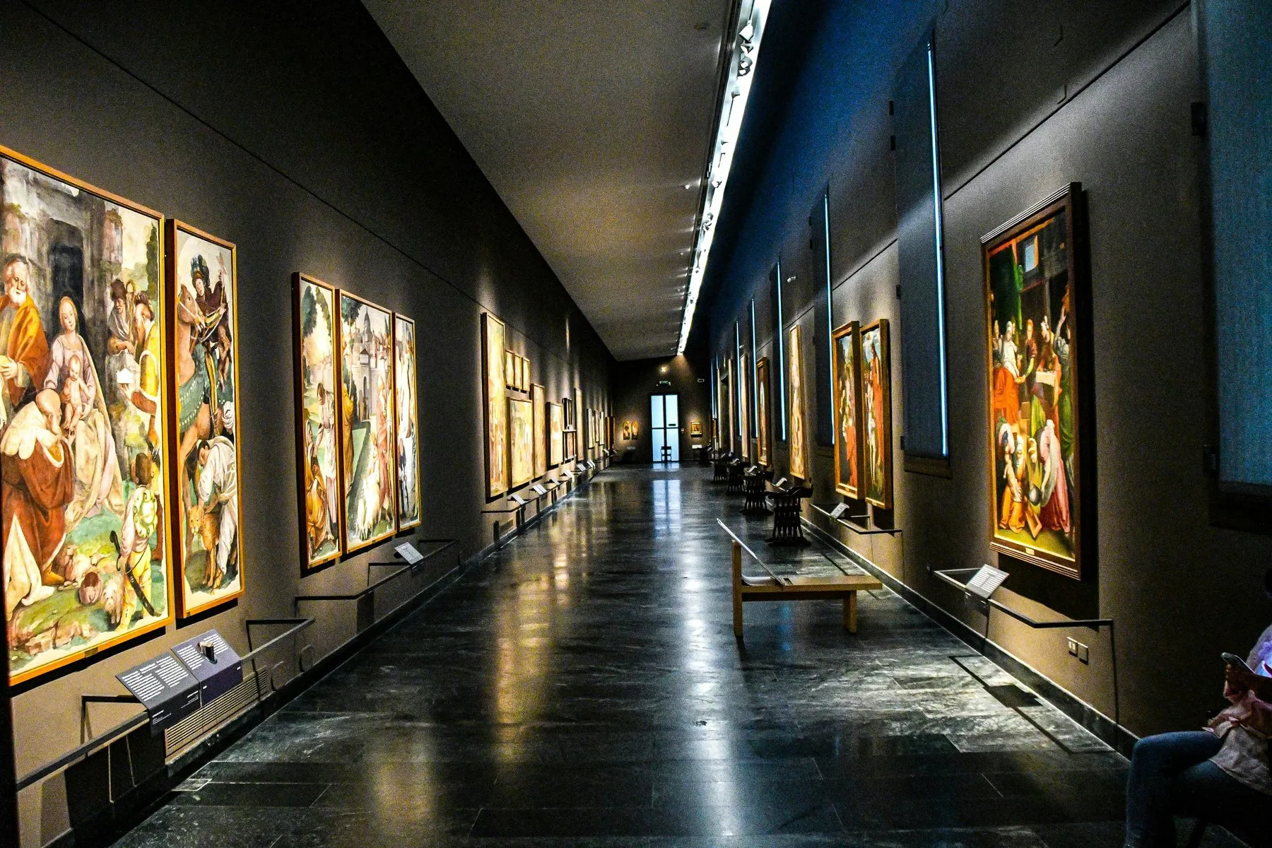En lang, mørk museumsgang med store malerier i gullrammer tett i tett langs begge vegger, og blankt gulv som speiler lyset fra spotlightene.