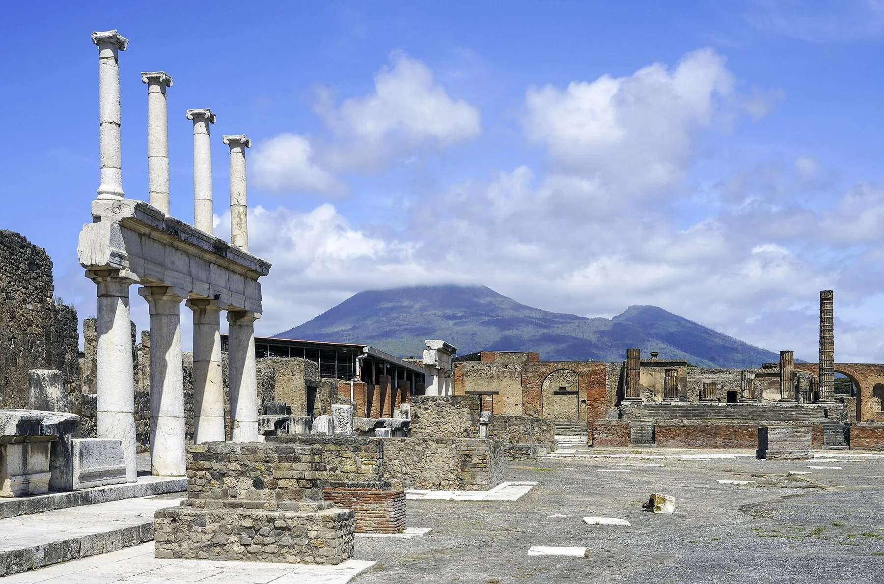 Utgravningsfeltet i Pompeii med en rad hvite marmorsøyler til venstre, murrester og steinlagt grunn utover, og Vesuv som reiser seg like bak den utgravde byen.