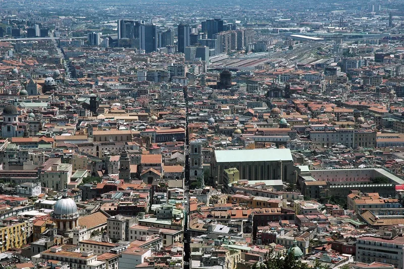 Sett ovenfra skjærer en knivrett, smal gate rett gjennom de tettpakkede takene i den historiske bydelen i Napoli, med kupler og kirketårn på begge sider og høyhus i det fjerne.