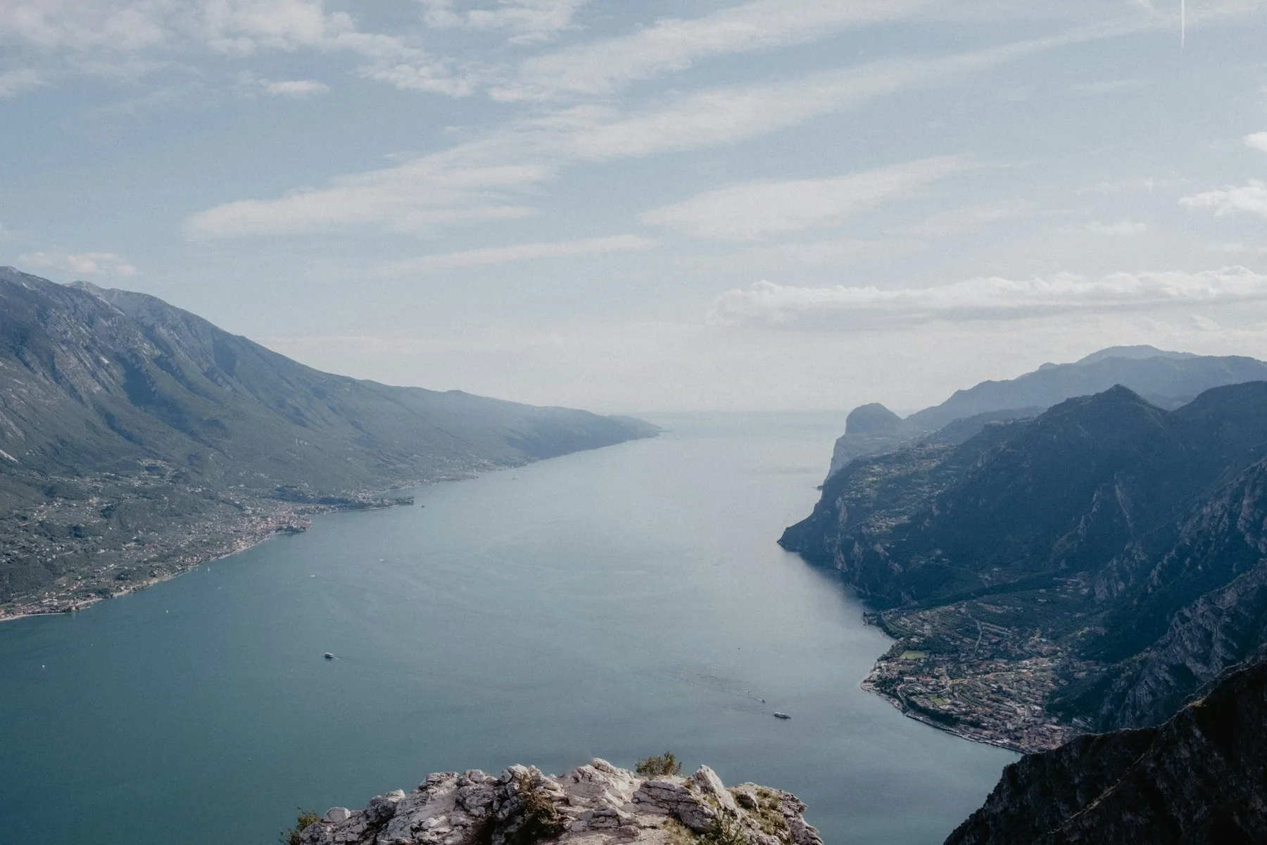 Utsikt fra en steinete fjellkant ned mot en lang innsjø som ligger klemt mellom bratte fjellsider, med disig horisont langt der framme.