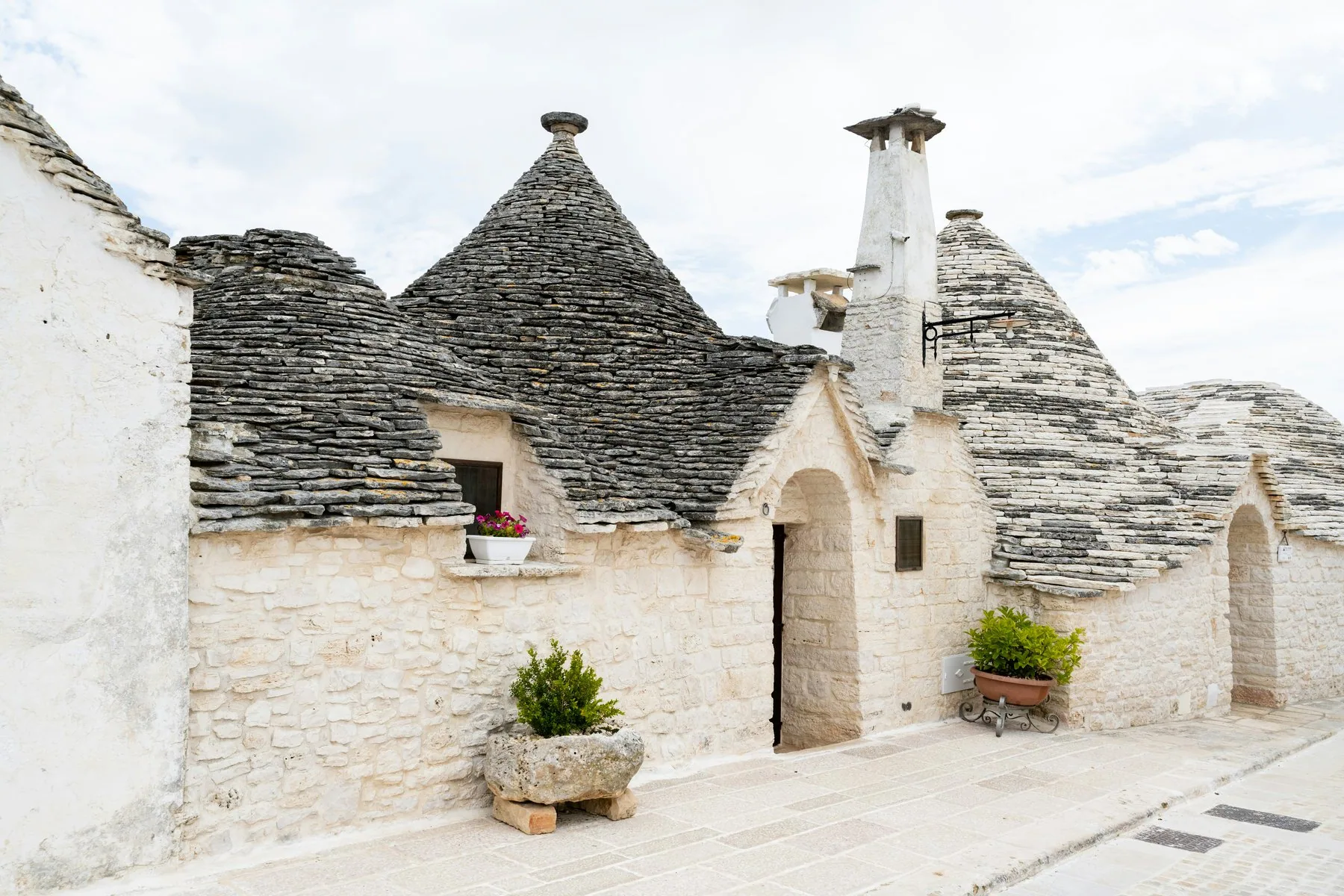 Hvitkalkede trulli-hus med kjegleformede tak av flate steinheller står på rekke langs en gate, med potteplanter ved dørene.