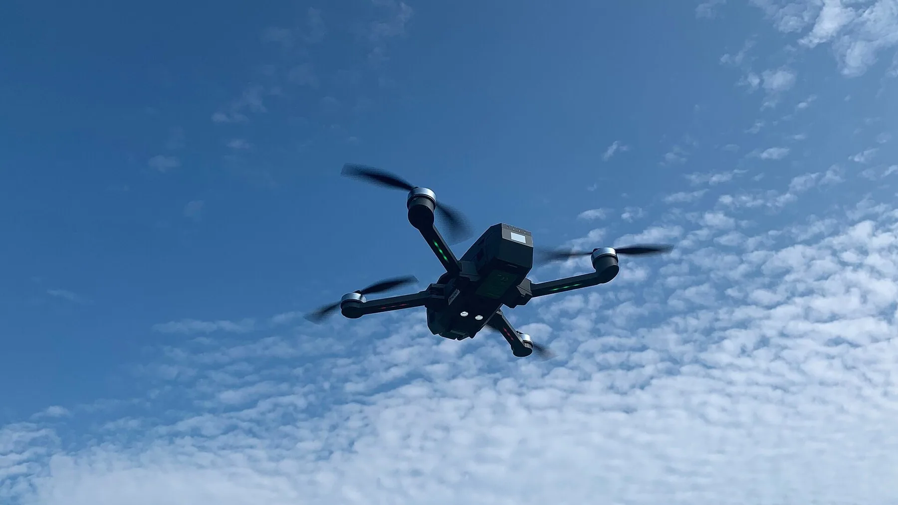 En mørk drone med fire roterende propeller og et kamera under kroppen henger i lufta, sett nedenfra mot blå himmel og et teppe av små hvite skyer.
