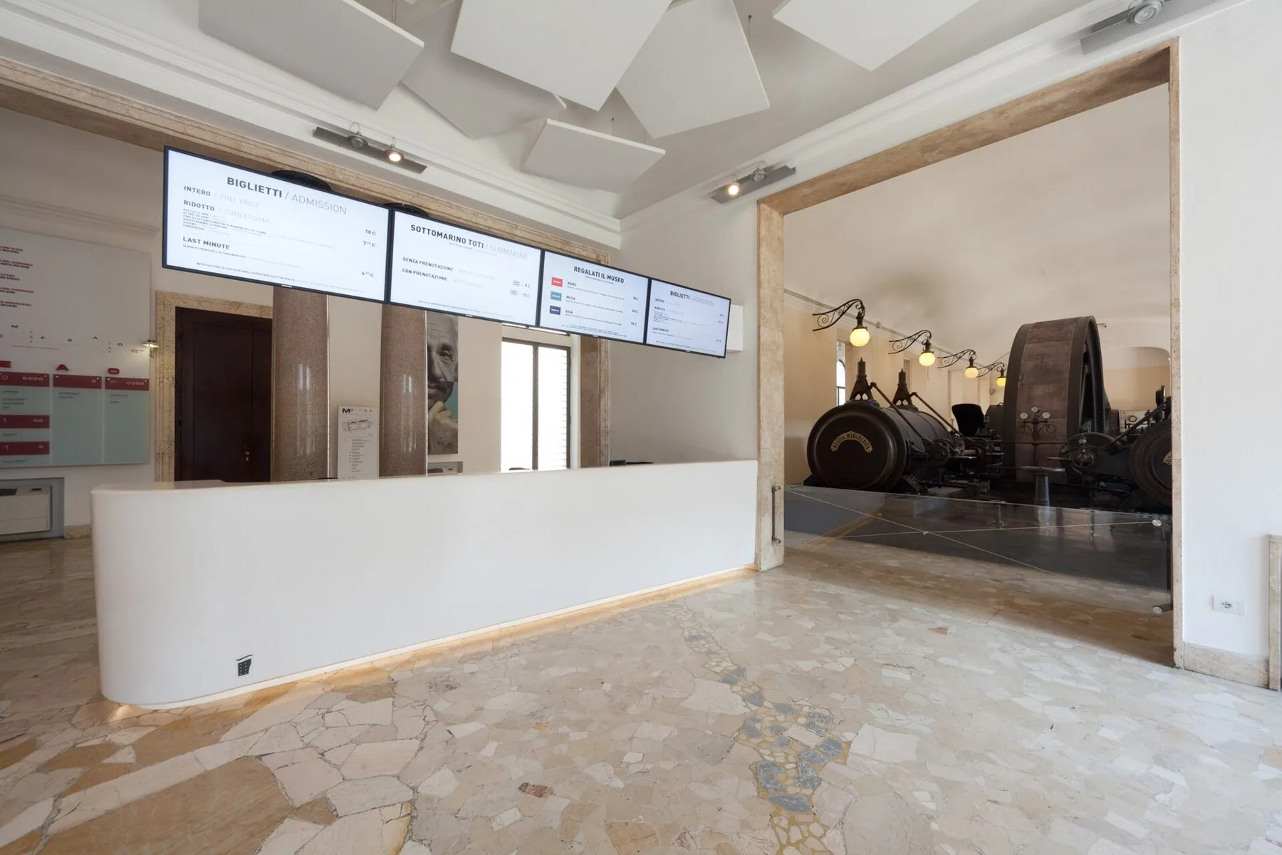 En lang hvit billettdisk i en museumsfoaje, med fire skjermer over disken som viser prisene for hel og redusert billett, og en gammel dampmaskin bak en glassvegg til høyre.