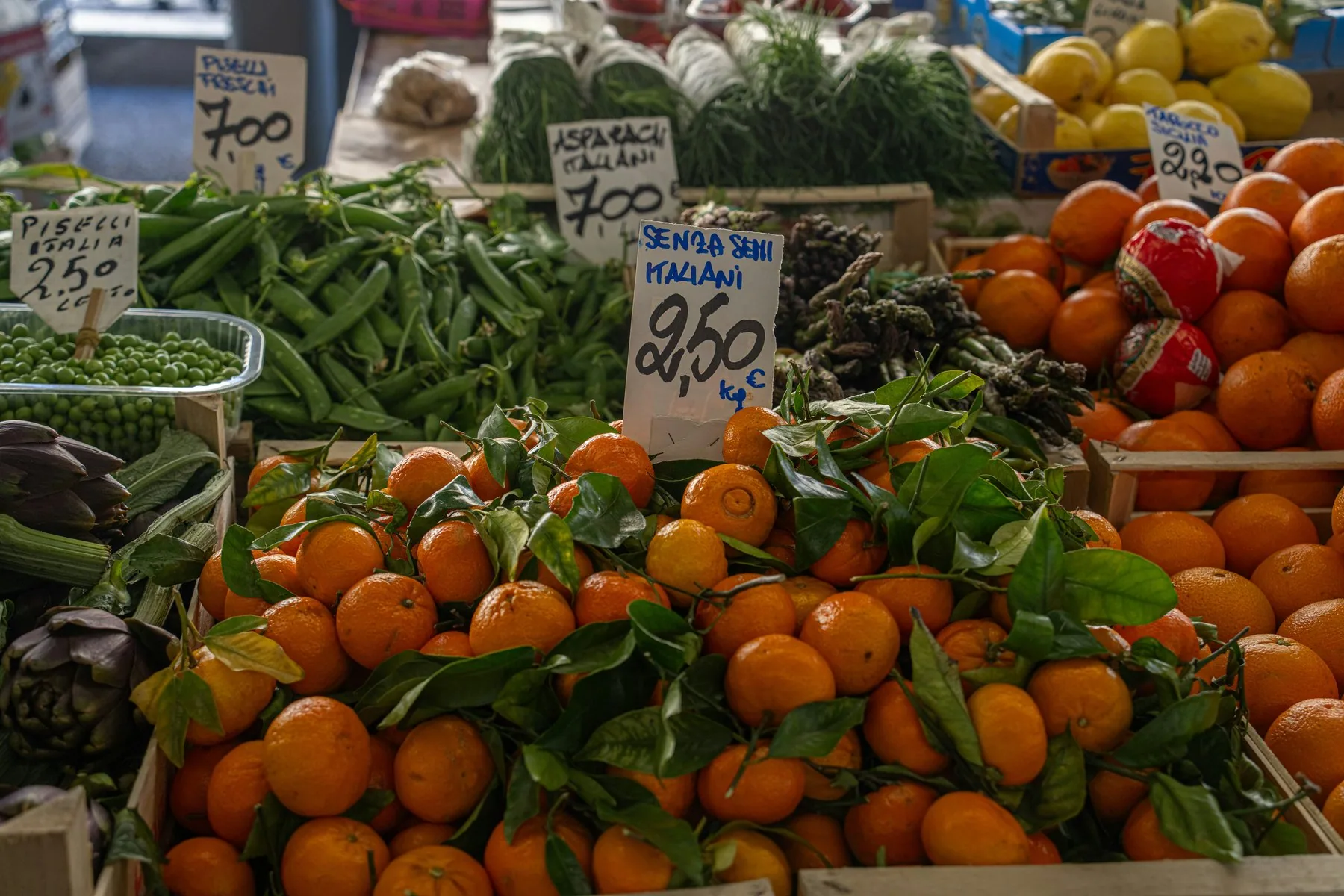 Appelsiner med blad, sitroner og grønnsaker ligger i kasser på en markedsbod, med håndskrevne prisskilt i euro stukket ned mellom varene.