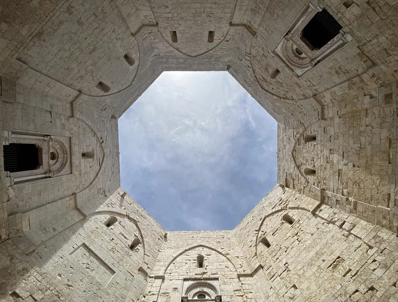 Den åttekantede gårdsplassen i Castel del Monte sett nedenfra, med kalksteinsvegger og vinduer rundt en åpning mot himmelen.