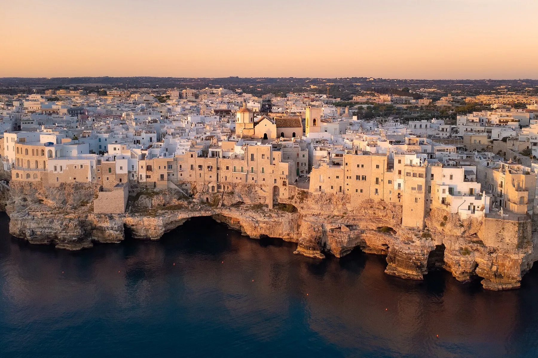 Den hvite gamlebyen i Polignano a Mare ligger helt ute på kanten av en kalksteinsklippe i morgenlys, med huler og grotter i klippeveggen og rolig sjø nedenfor.