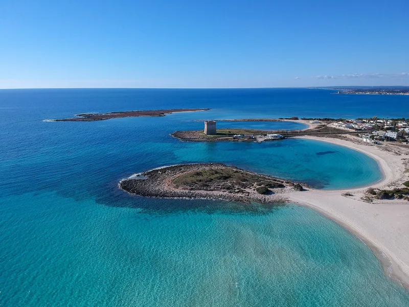 Små lave øyer og et gammelt vakttårn ligger i turkist, grunt vann utenfor en lang, hvit sandstrand, sett fra lufta.