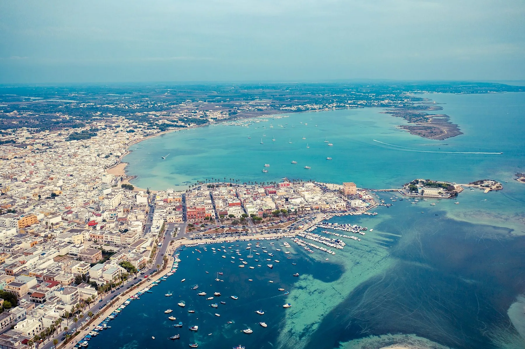 Porto Cesareo sett fra lufta, med småbåthavn, lave hus langs strandkanten og grunt, turkist vann med små øyer.