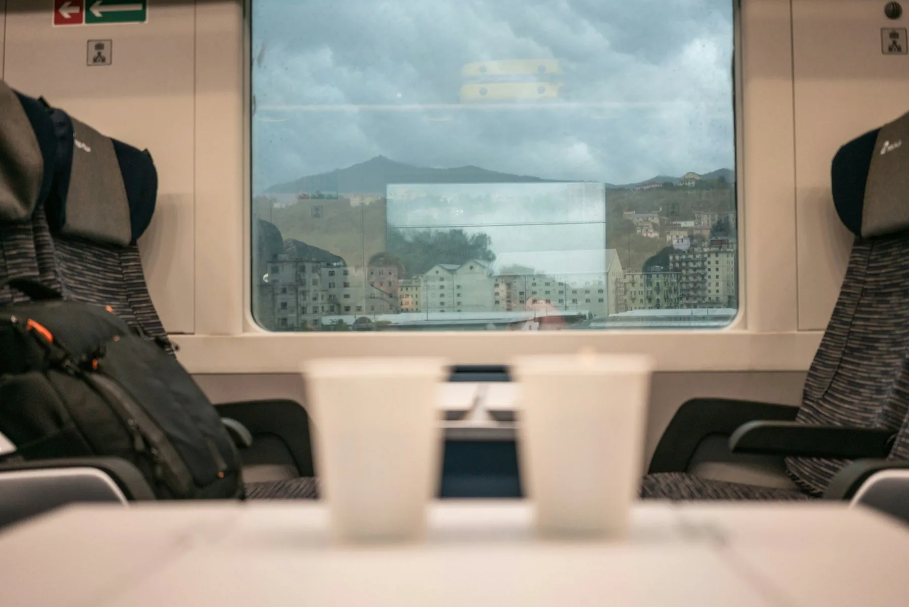 Utsikt gjennom et togvindu mot en by som klatrer oppover en kystskråning, med to hvite papirkopper uskarpt på bordet mellom setene i forgrunnen.