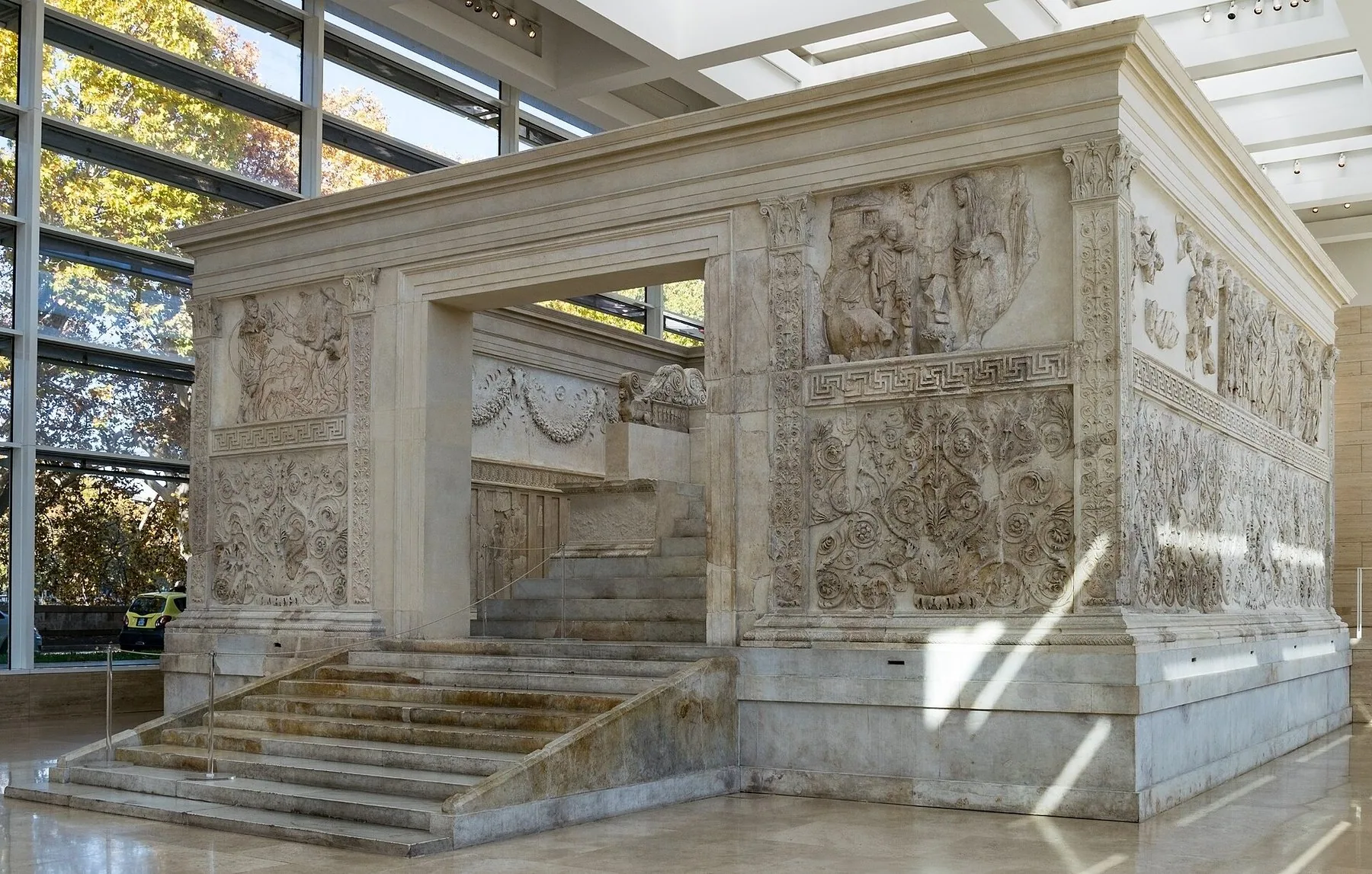 Ara Pacis Augustae: et marmoralter med trapp opp til åpningen og vegger dekket av relieffer med figurer og rankeornamentikk, inne i en lys museumshall med glassvegger.